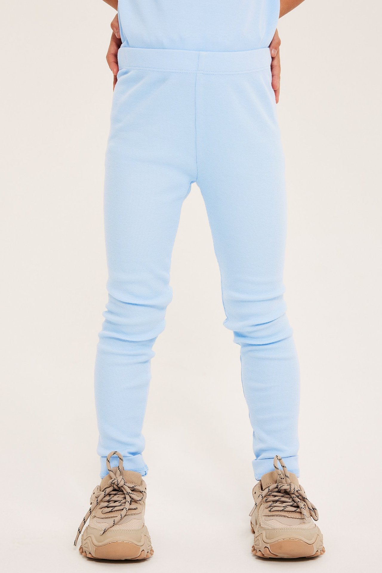 Frontvy av ett barn i ljusblå ribbade bomullsleggings, matchande topp och beige sneakers.