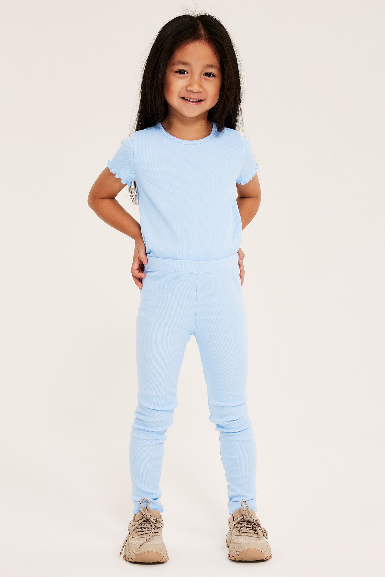 Widok z przodu dziecka w jasnoniebieskich prążkowanych legginsach, pasującym t-shircie i beżowych sneakersach.