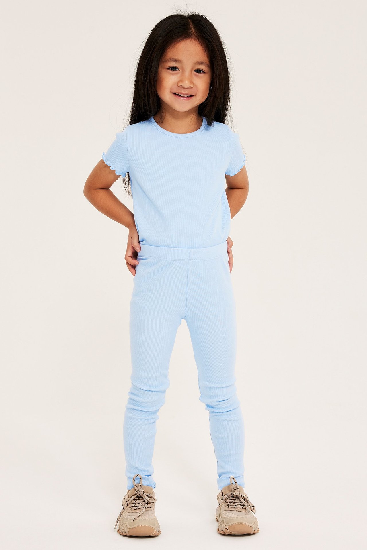 Frontvy av ett barn i ljusblå ribbade leggings, en matchande t-shirt och beiga sneakers.