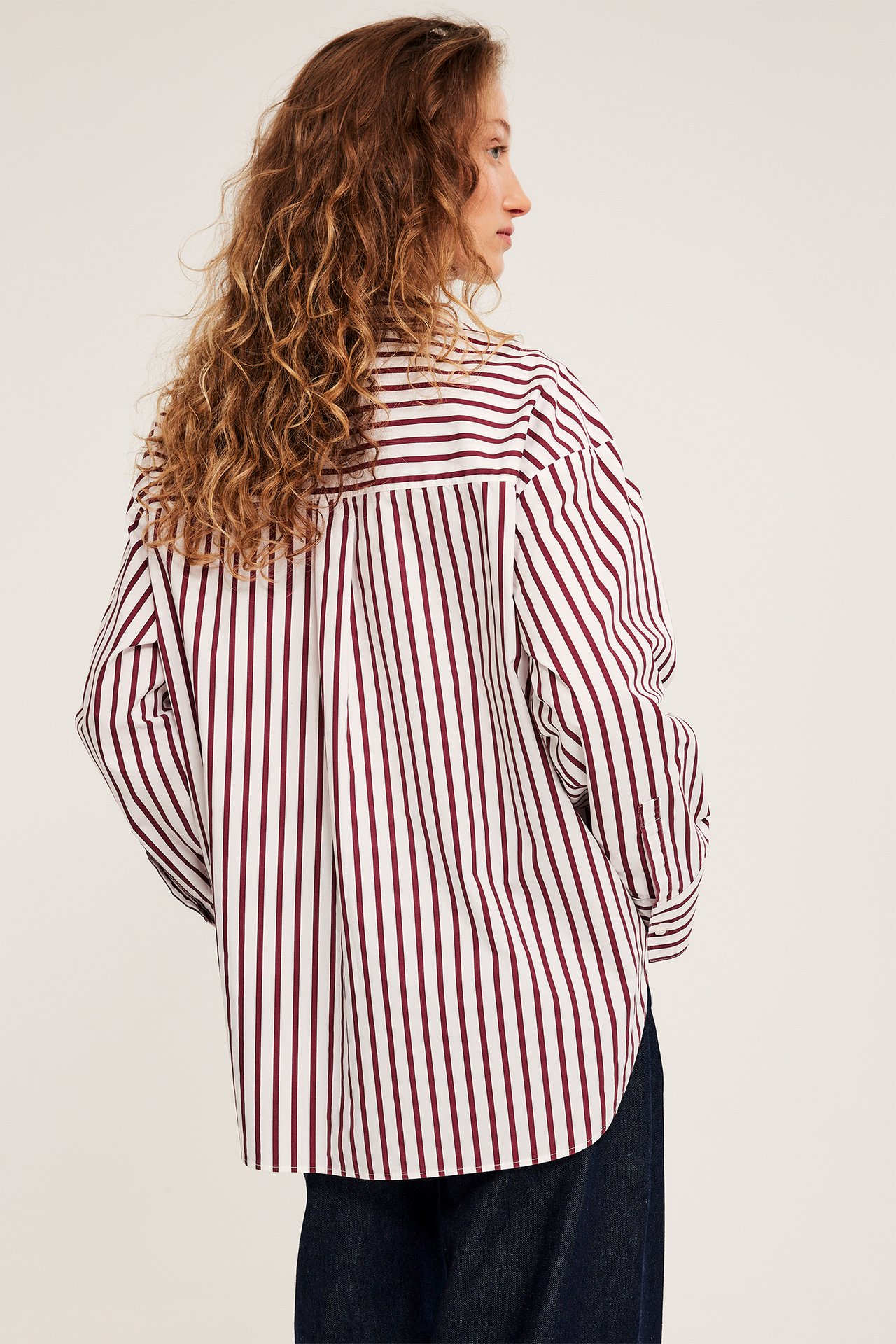 Naisen takakuva: luonnonvalkoinen ja viininpunainen raidallinen puuvillapopliinipaita tummien farkkujen päällä.