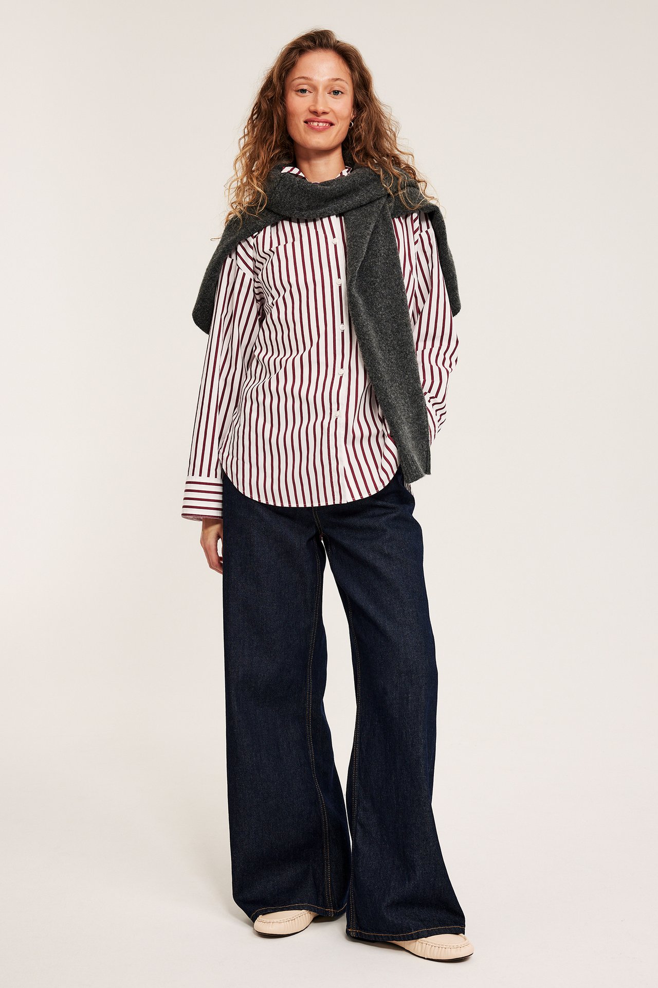 Edestäpäin: nainen yllään luonnonvalkoinen, raidallinen popliinipaita, tummansiniset leveälahkeiset farkut ja harmaa neule.