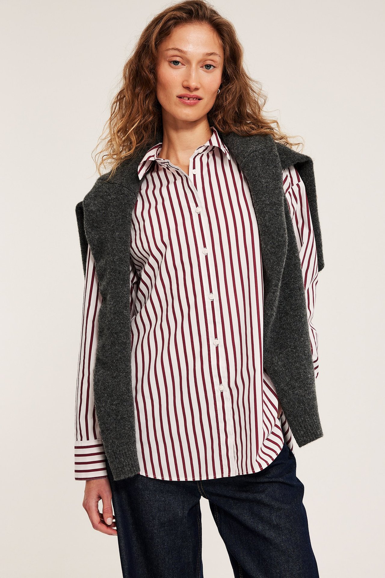 Edestäpäin, nainen yllään offwhite ja viininpunainen raidallinen popliinipaita, tummanharmaa neule olkapäillä ja tummansiniset farkut.