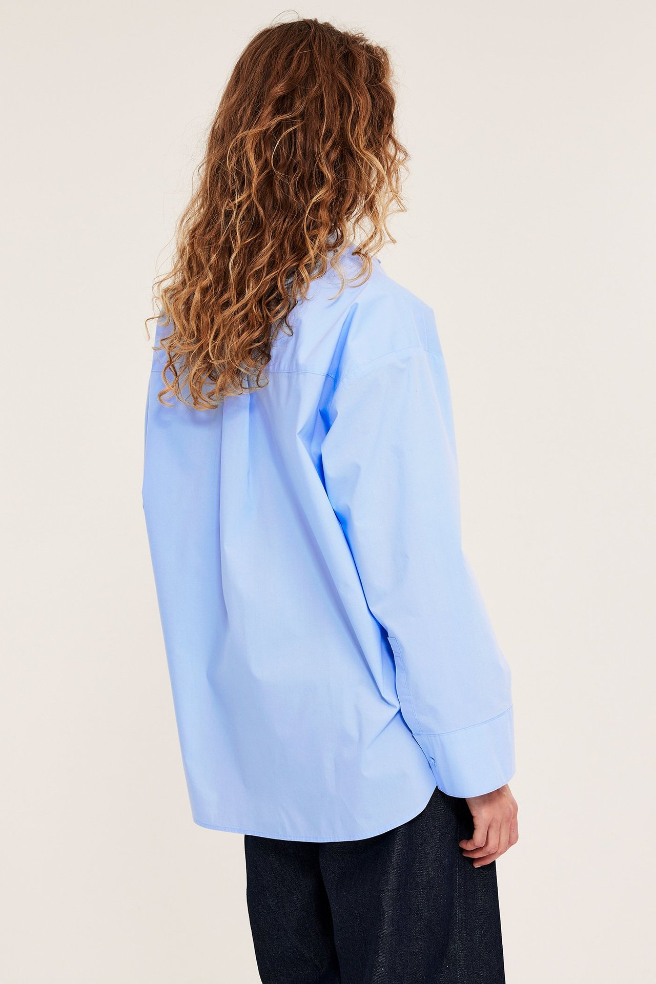 Naisen takaapäin, yllään väljä vaaleansininen puuvillapopliinipaita pitkillä hihoilla ja tummat housut.