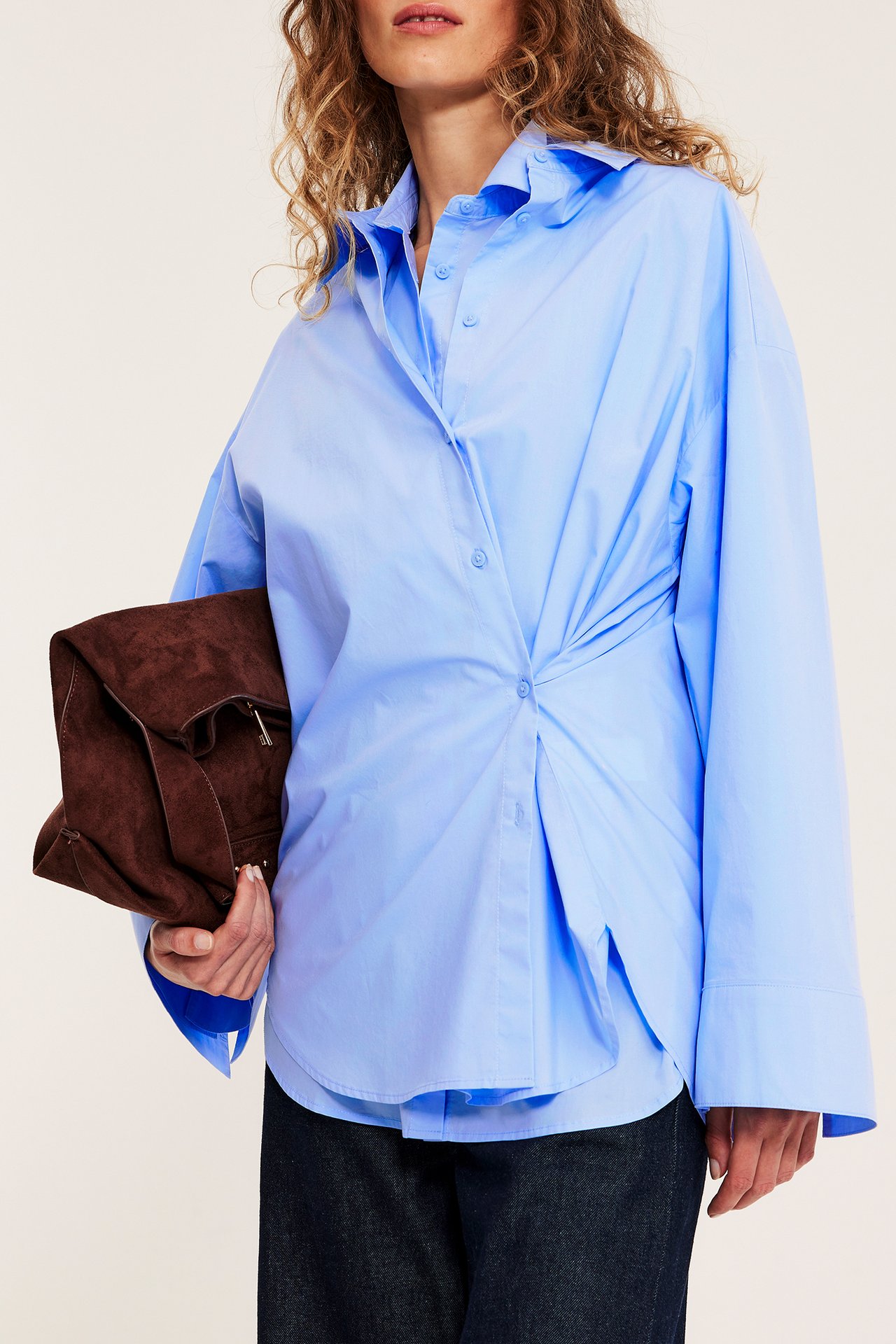 Edestä nainen yllään oversize vaaleansininen popliinipaita kierretysti napitettuna, tummat housut, pitää ruskeaa mokkalaukkua.