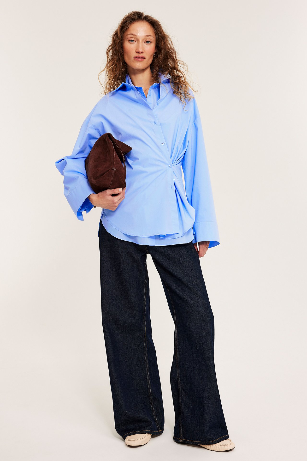 Edestäpäin: Nainen vaaleansinisessä popliinipaidassa sivusolmuilla, tummansinisissä farkuissa, pitäen ruskeaa clutch-laukkua.