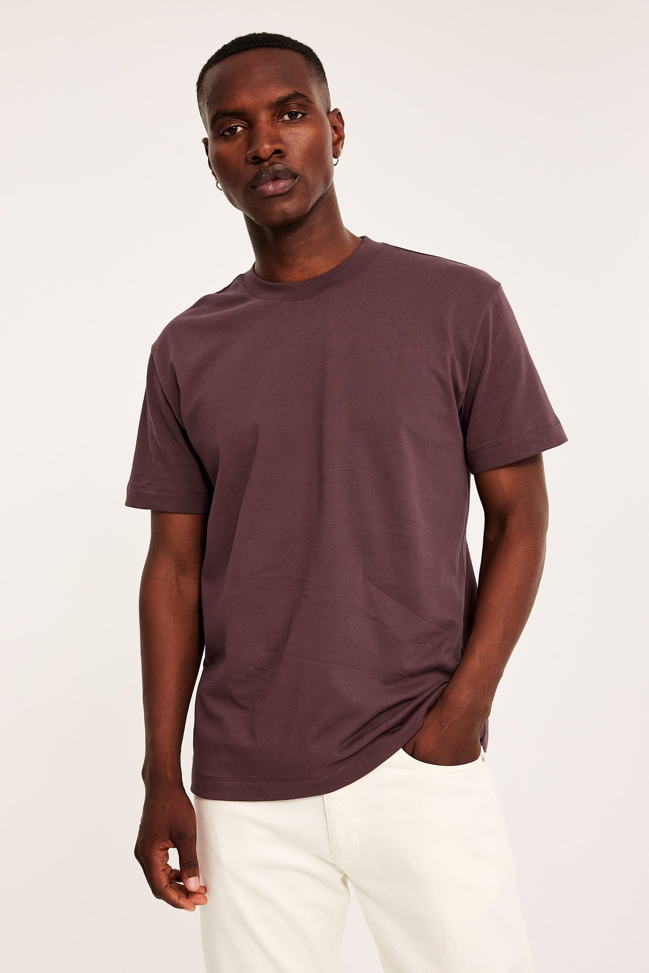 Framifrån: man i en mörkröd, lös passform bomulls t-shirt och vita byxor, med handen i fickan.