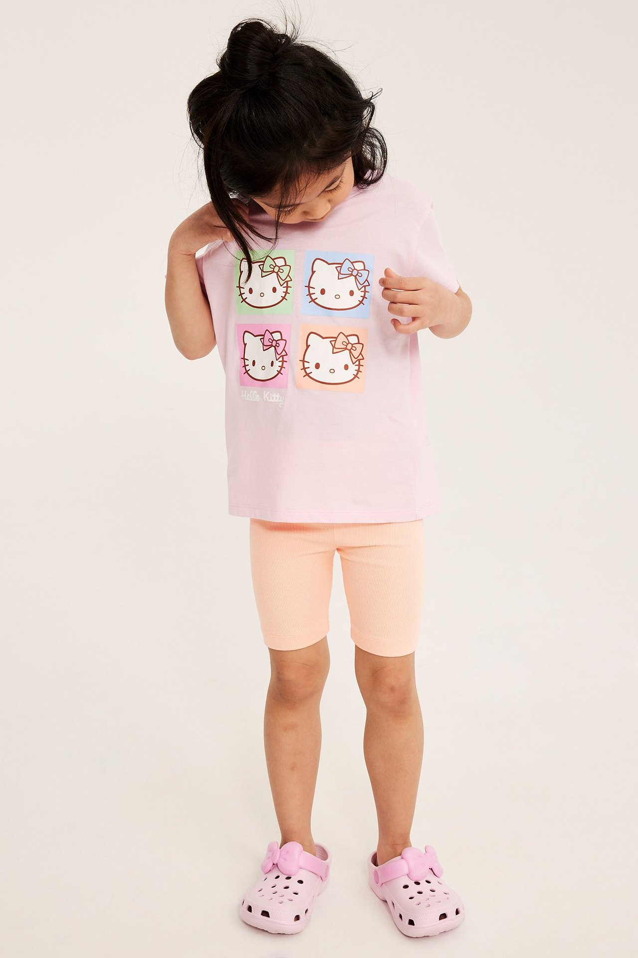 Edestä: Lapsi pukeutuu vaaleanoransseihin ribbattuihin pyöräilyhousuihin, vaaleanpunaiseen Hello Kitty t-paitaan ja vaaleanpunaisiin pistokkaisiin.