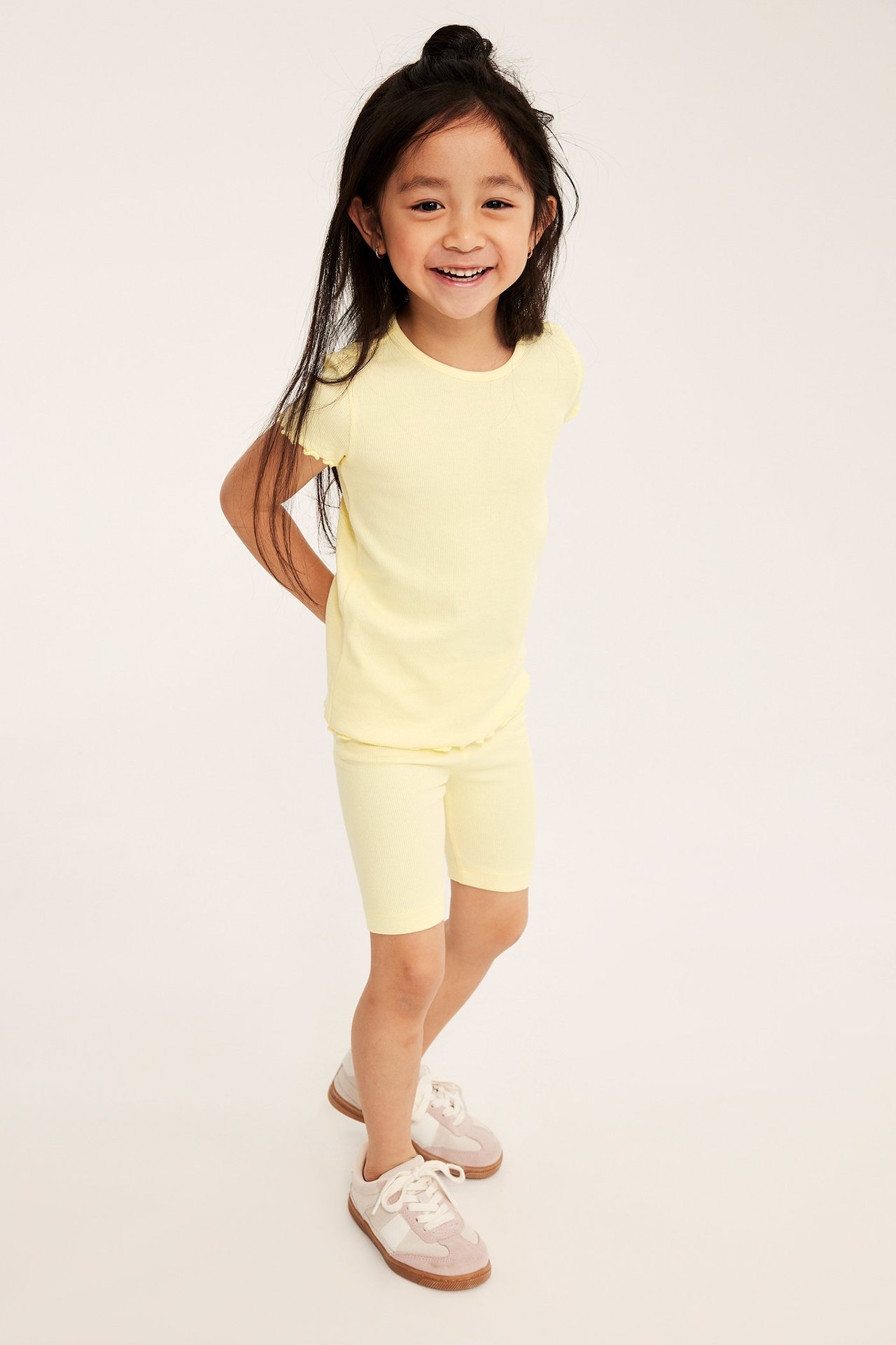 Frontvisning av et barn i lysegul ribbet sykkelshorts, matchende t-skjorte og beige/rosa sneakers.