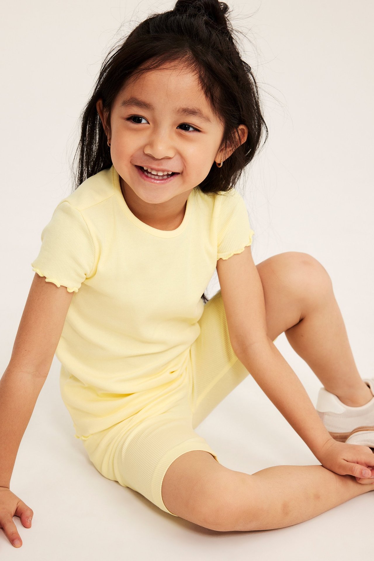 Edestäpäin: Hymyilevä lapsi vaaleankeltaisissa ribbireisihousuissa ja yhteensopivassa lyhythihaisessa paidassa.