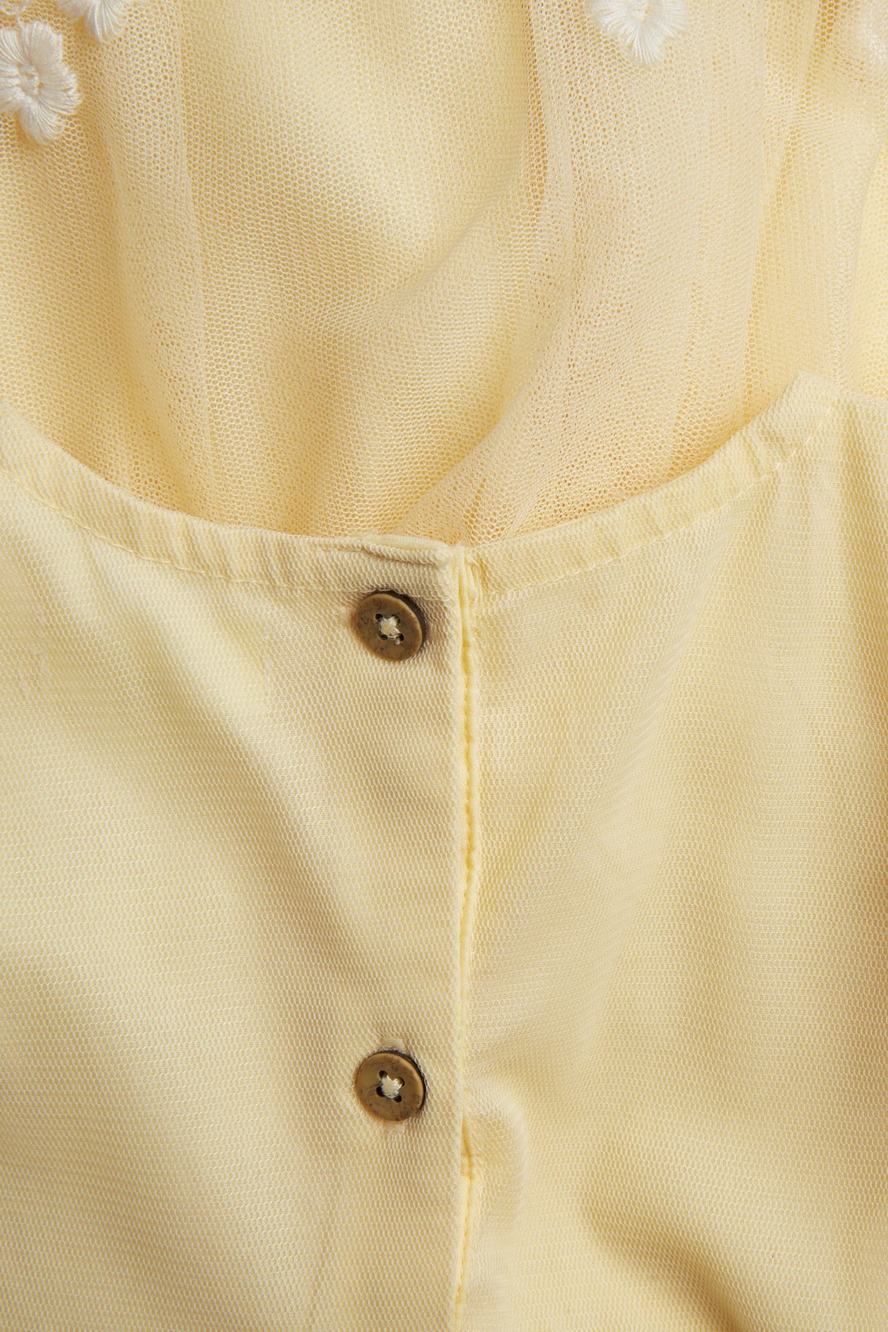 Lähikuva keltaisesta verkkomekosta, jossa valkoisia kukkakirjontaa ja kaksi ruskeaa nappia.