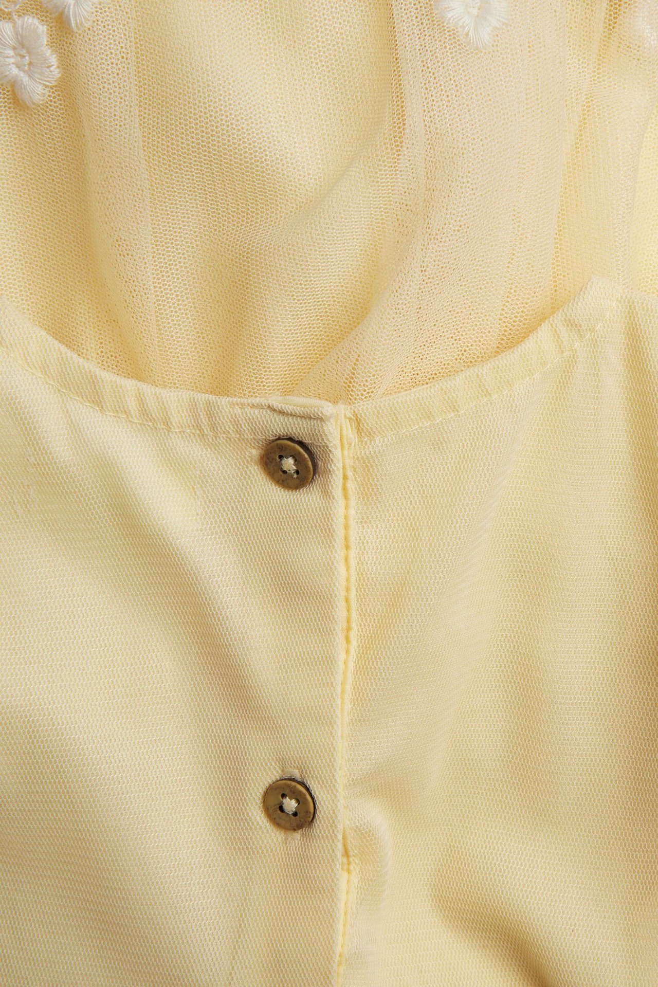 Zbliżenie żółtej sukienki z siateczki z białym haftem kwiatowym i dwoma brązowymi guzikami.