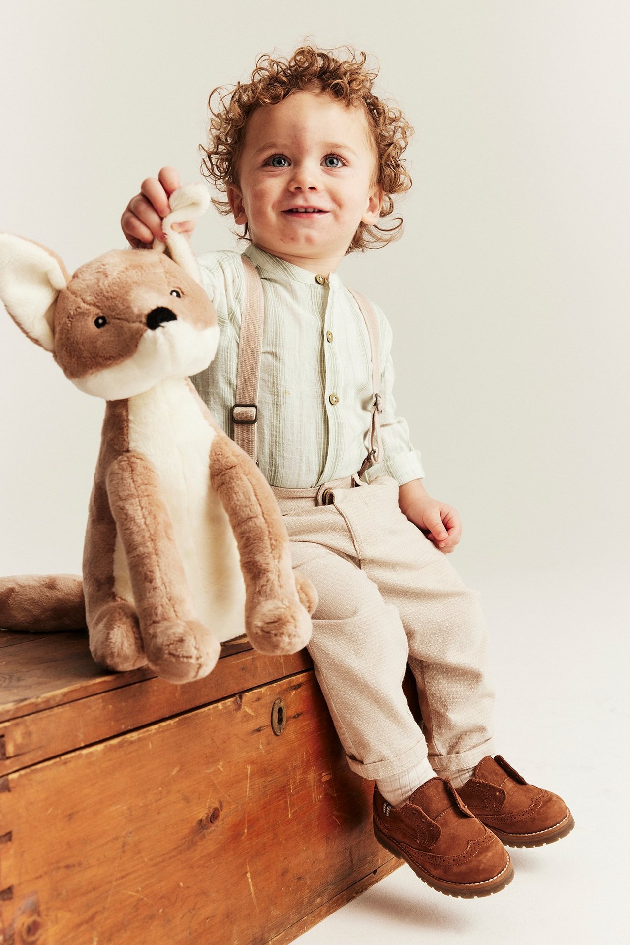 Framifrån, en baby i ljusgrön randig långärmad skjortbody, beige byxor med hängslen och bruna skor, håller en känguruleksak.