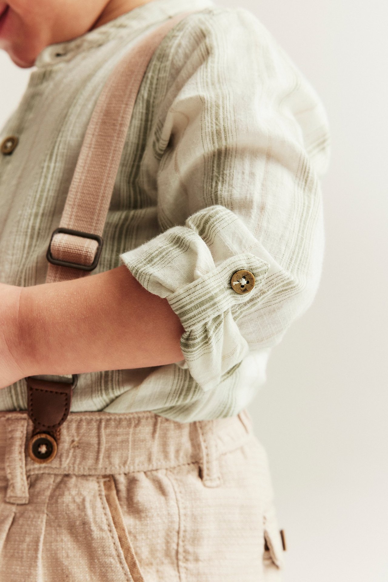 Zbliżenie na dziecko w jasnozielone paski body koszulowym z podwiniętymi rękawami, beżowych spodniach i różowych szelkach.