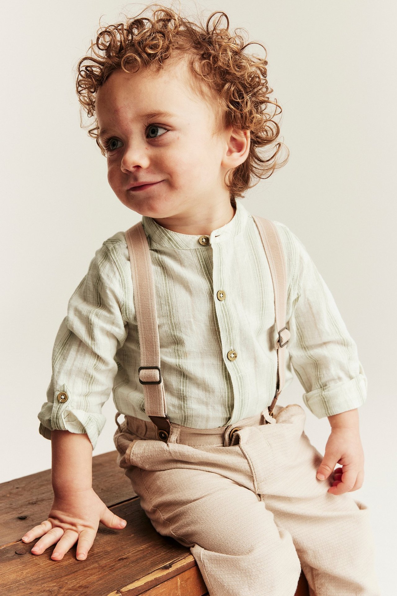 Z przodu: niemowlę w jasnozielonym pasiastym body, beżowych spodniach i szelkach.