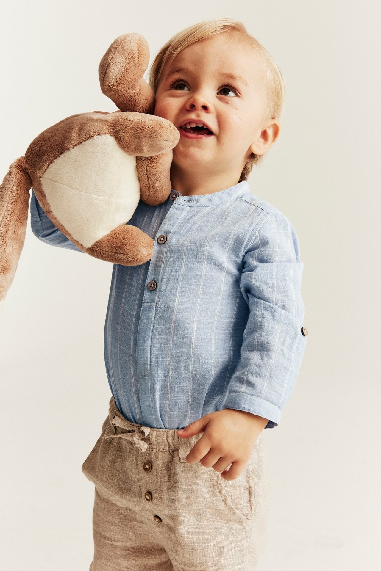 Babygutt har på seg en blåstripet langermet skjortebody og beige shorts, holder en brun koseleke.
