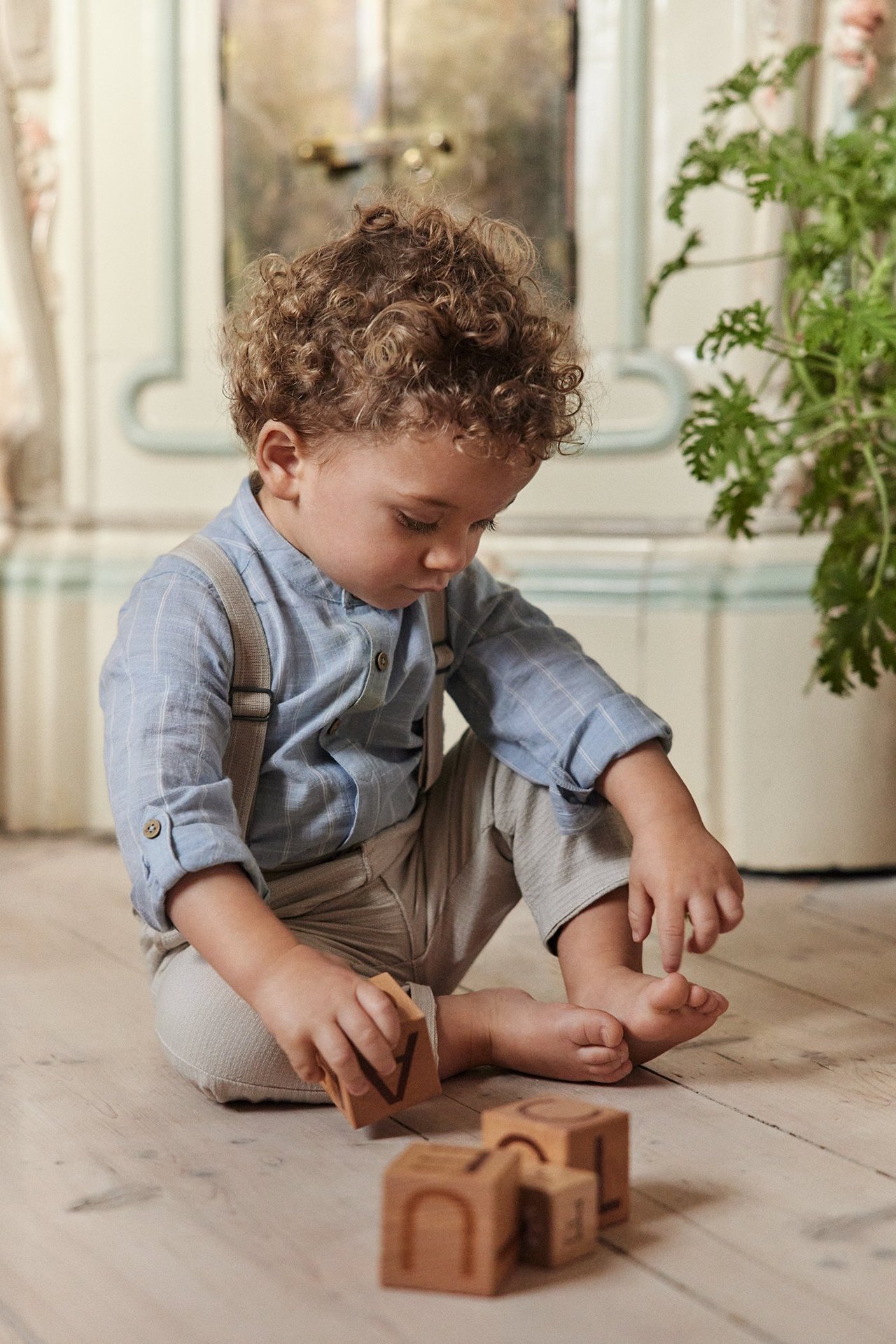 Widok z przodu: Dziecko w jasnoniebieskim pasiastym body koszulowym ze stójką, beżowych spodniach, szelkach.