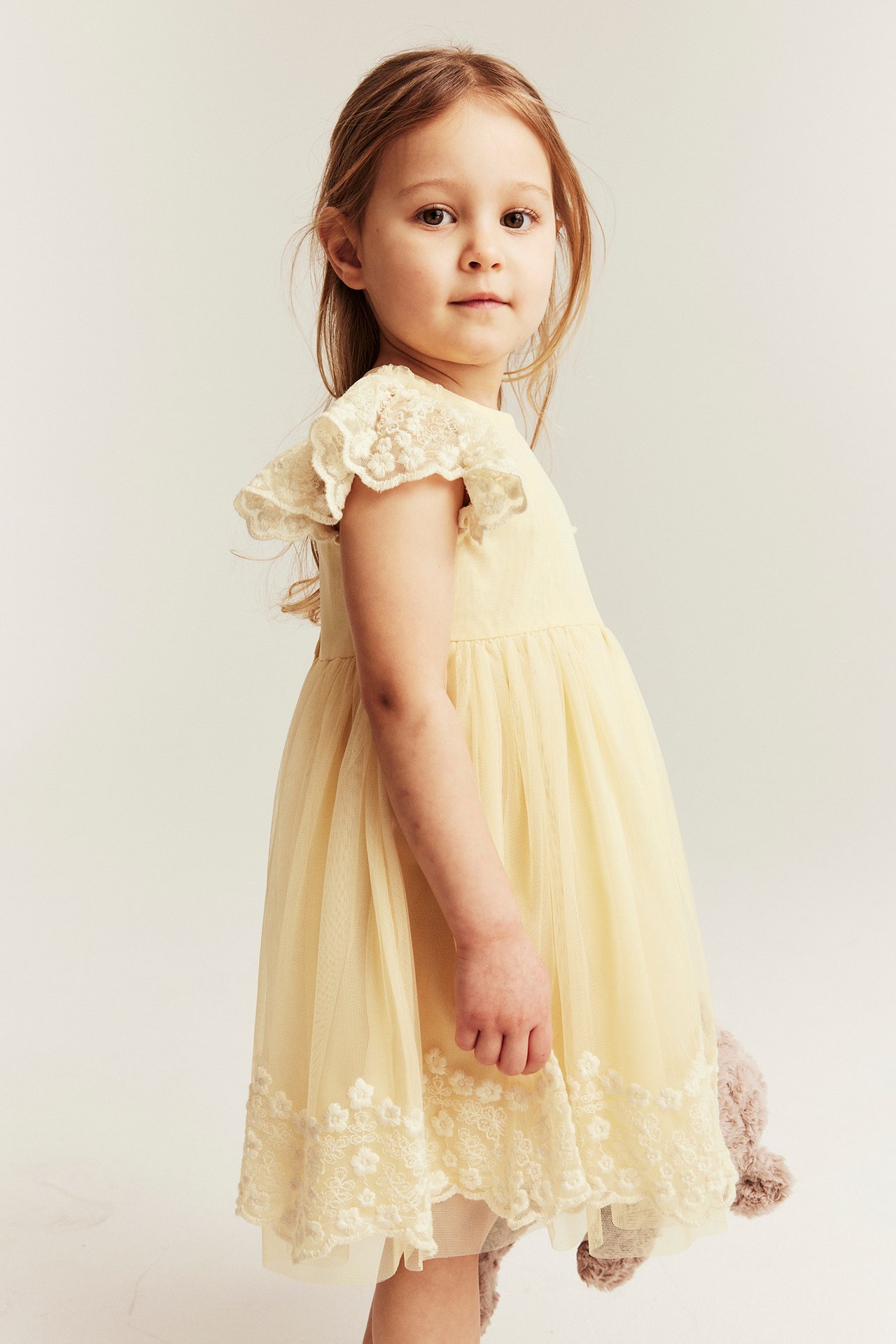 Forfra, en jente i gul meshkjole med broderte ermer og kant, holder et kosedyr.