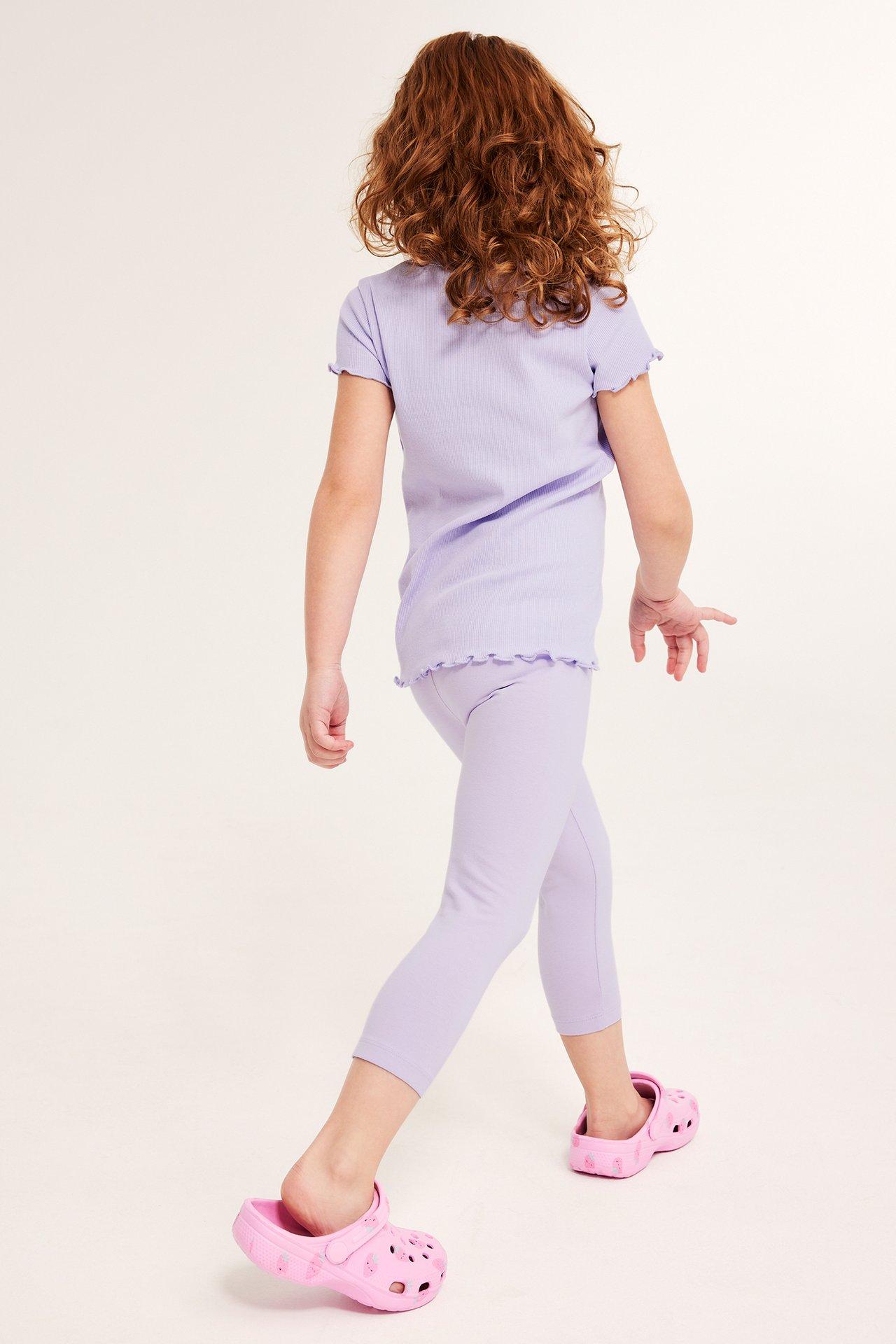 Bakfra et barn iført lys lilla ribbestrikket capri-leggings, matchende topp og rosa crocs.