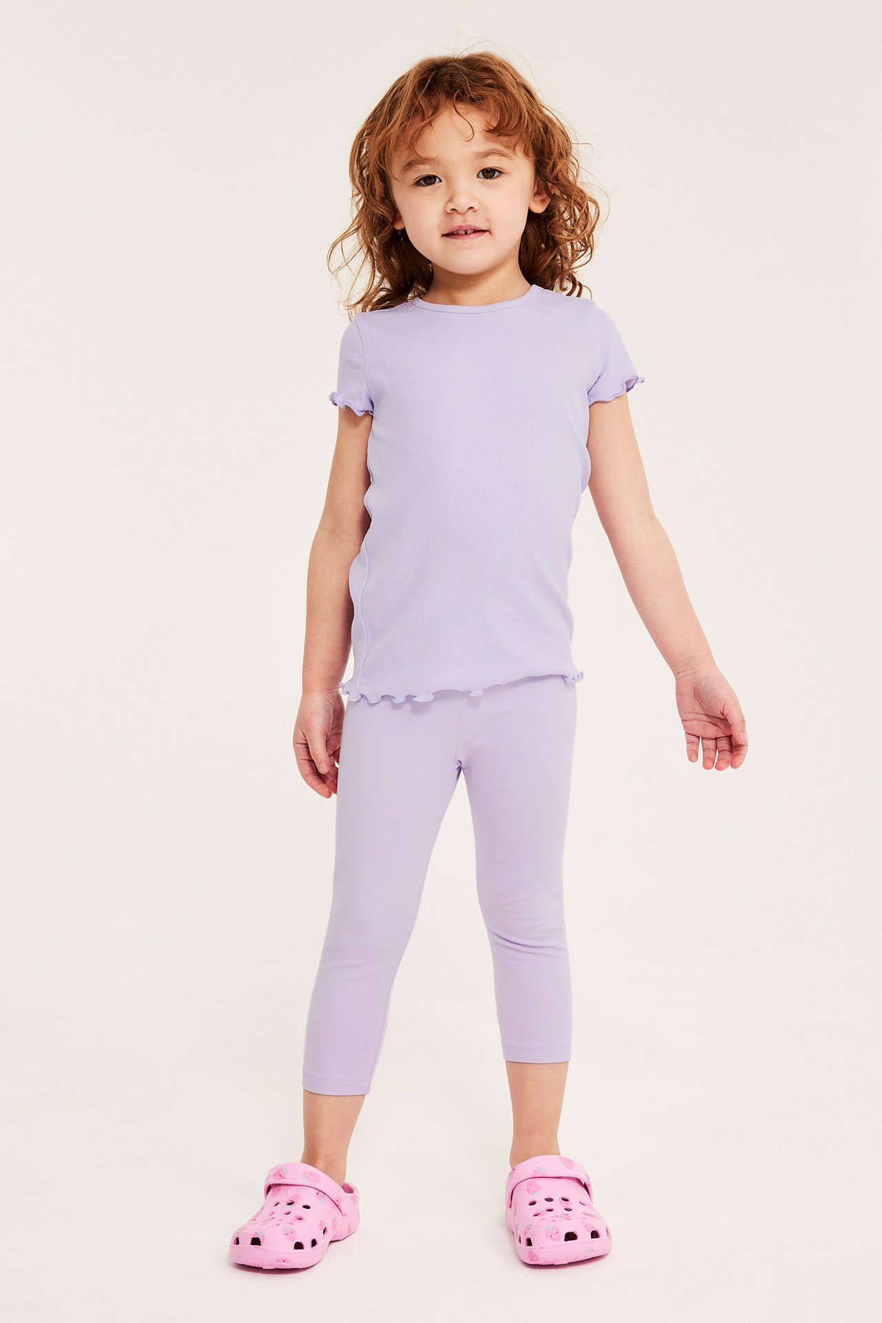 Framifrån, ett barn i ljuslila 3/4-leggings, matchande volangtopp och rosa tofflor.