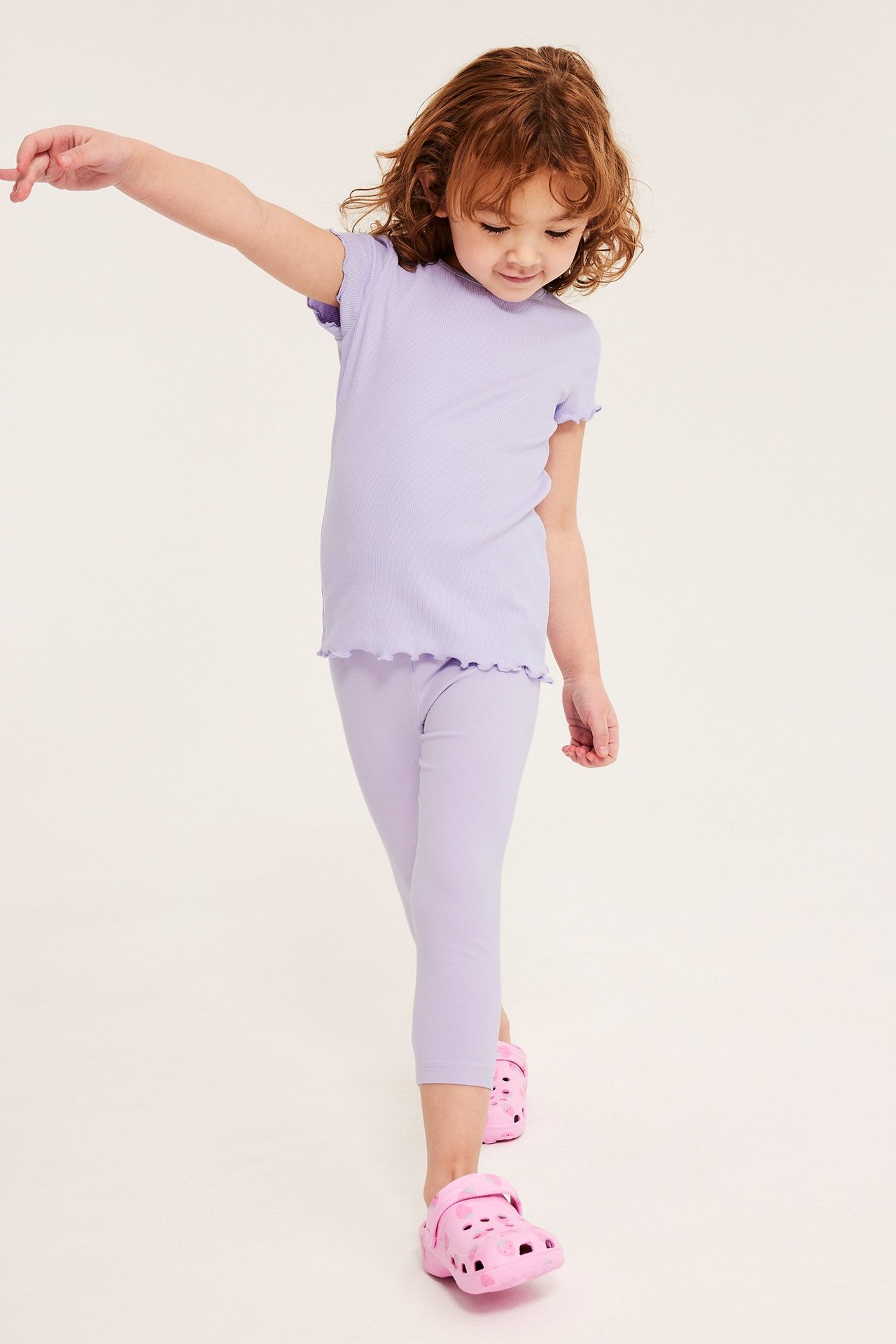 Framifrån: Barn i ljuslila enfärgade caprileggings, matchande t-shirt med volanger, rosa tofflor.