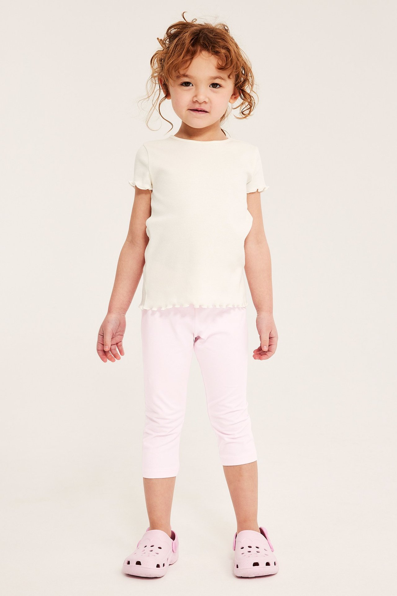 Framifrån, ett barn i ljusrosa caprileggings, en vit ribbad t-shirt och rosa tofflor.