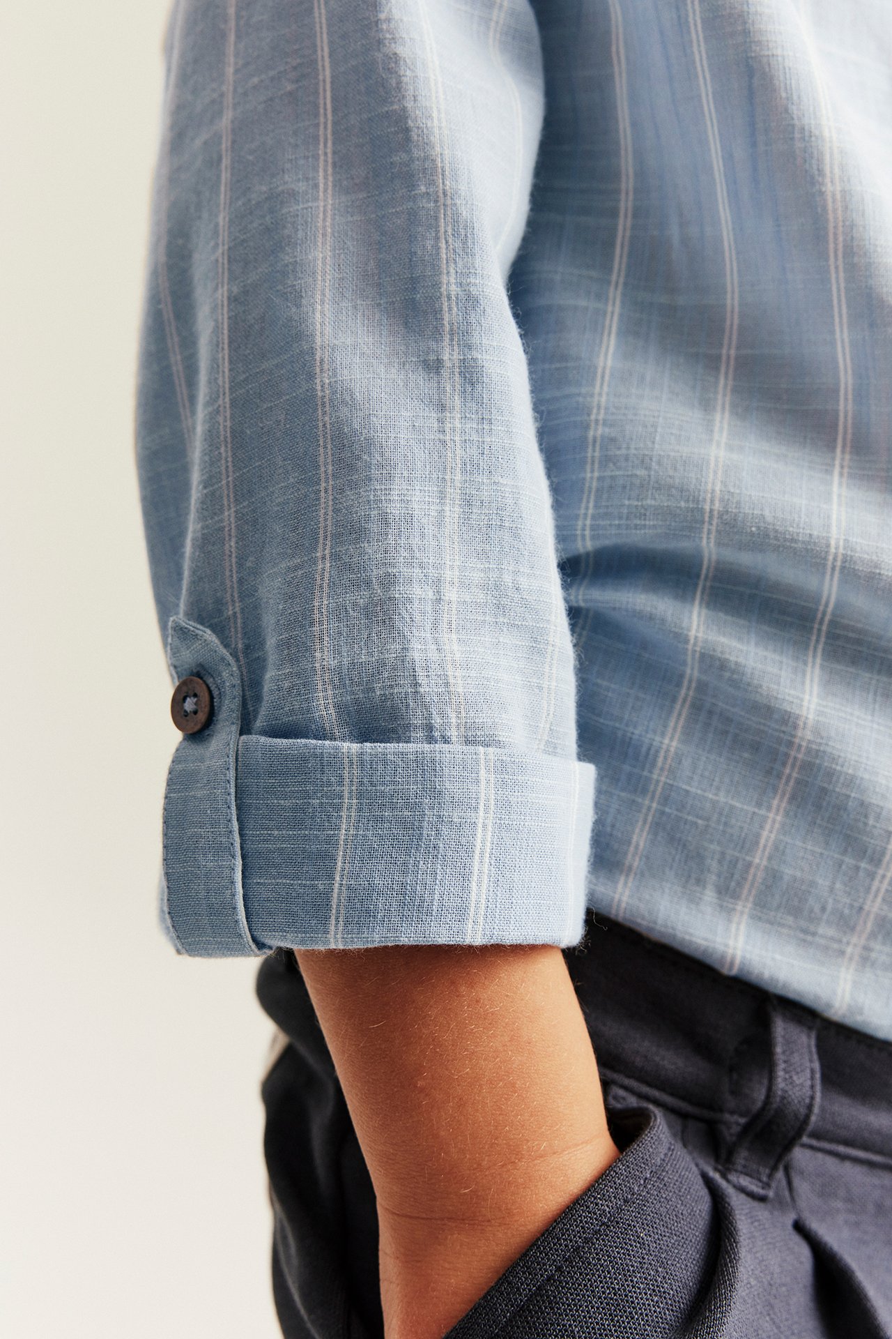 Närbild på ett barns arm i en blårandig bomullsskjorta med upprullad ärm, över mörka byxor.