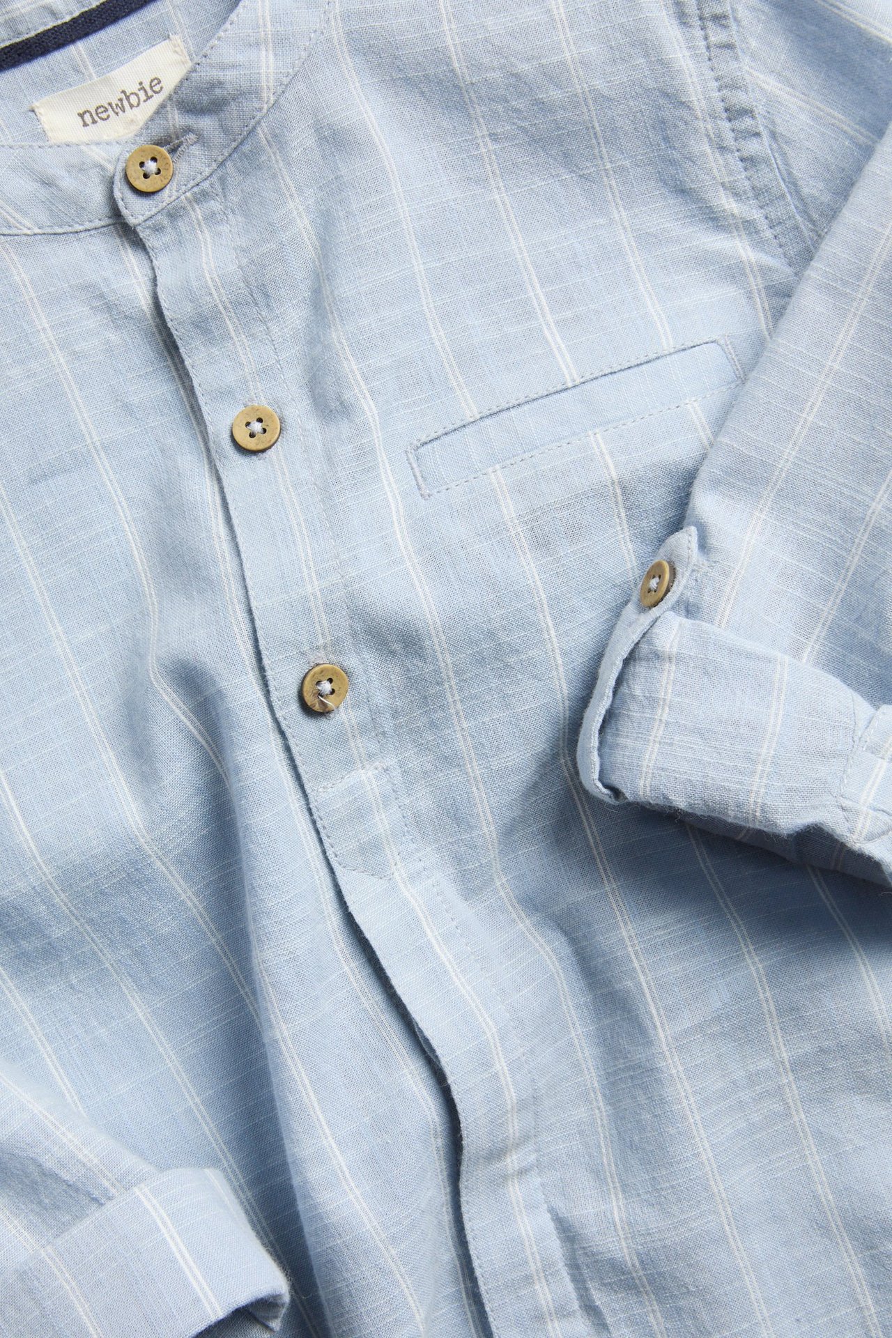 Närbild på en barnskjorta i ljusblå randig bomull med murarkrage, träknappar och upprullade ärmar.