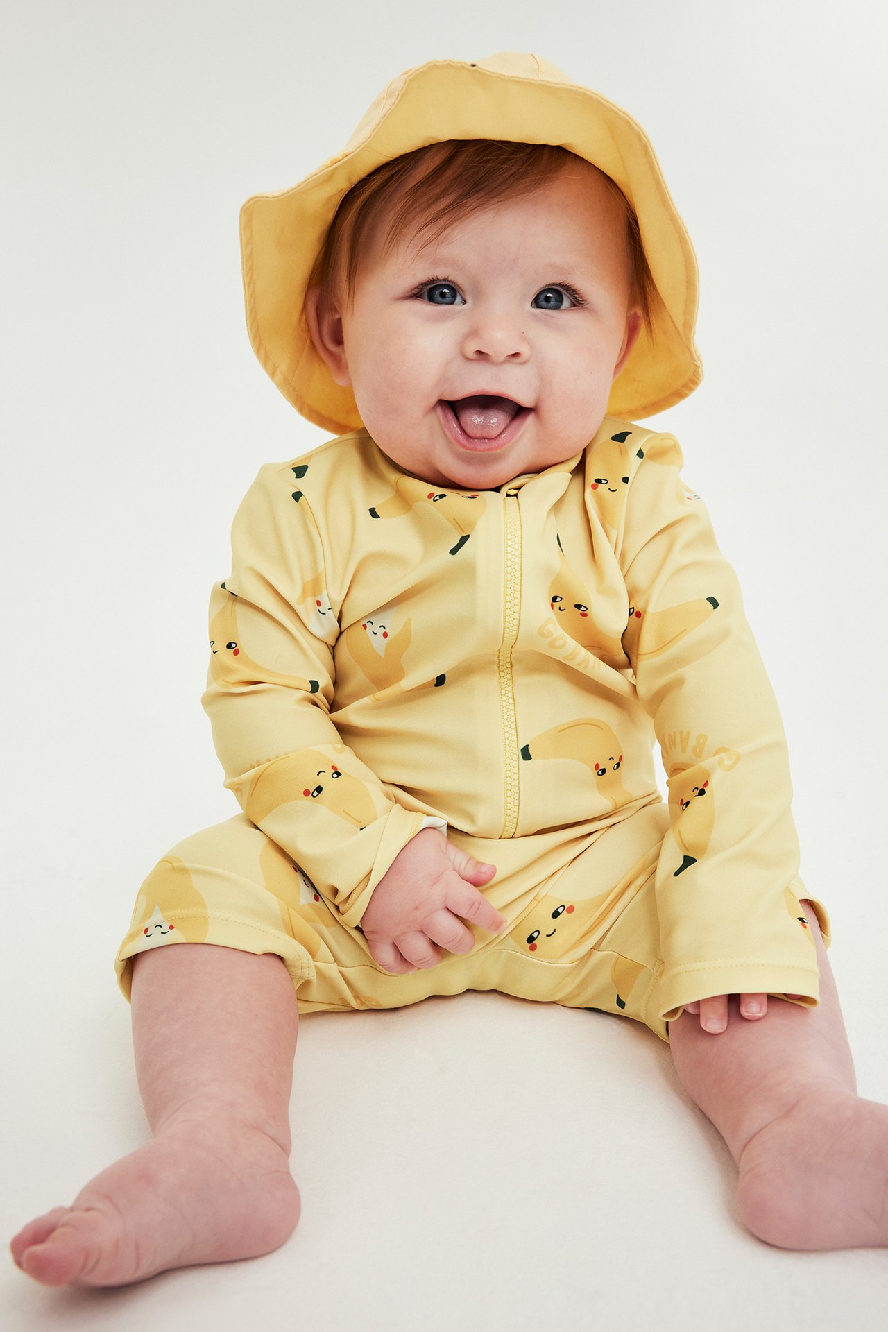 Smilende baby i gul UV-drakt med bananprint, glidelås foran og matchende solhatt.