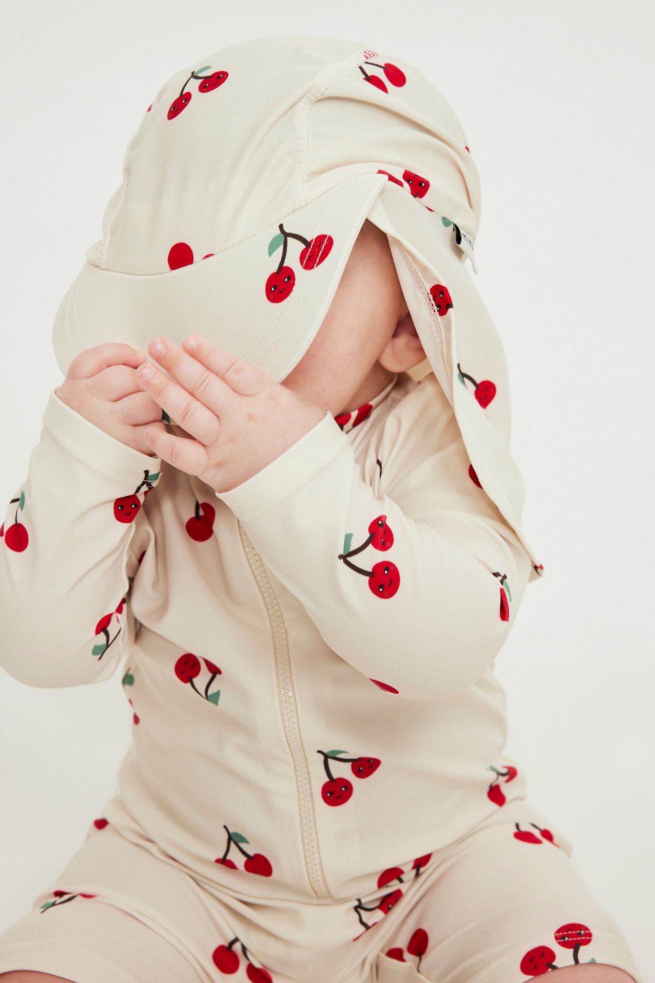 Nærbilde av baby i lys beige UV-drakt med kirsebærmønster og glidelås foran.