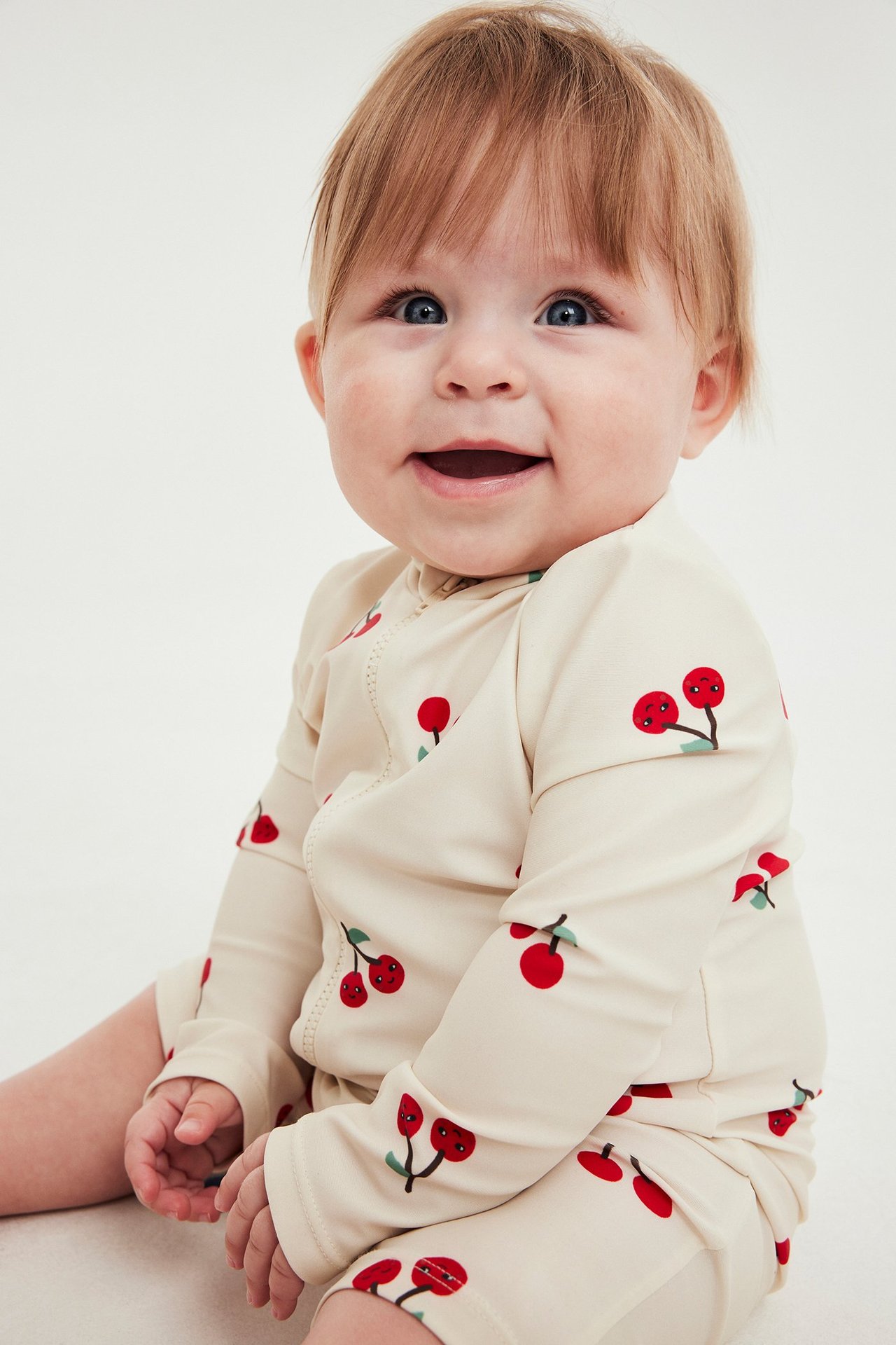 Lähikuva edestä hymyilevästä vauvasta, yllään vaalea beige UV-asu kirsikkakuviolla ja vetoketjulla edessä.