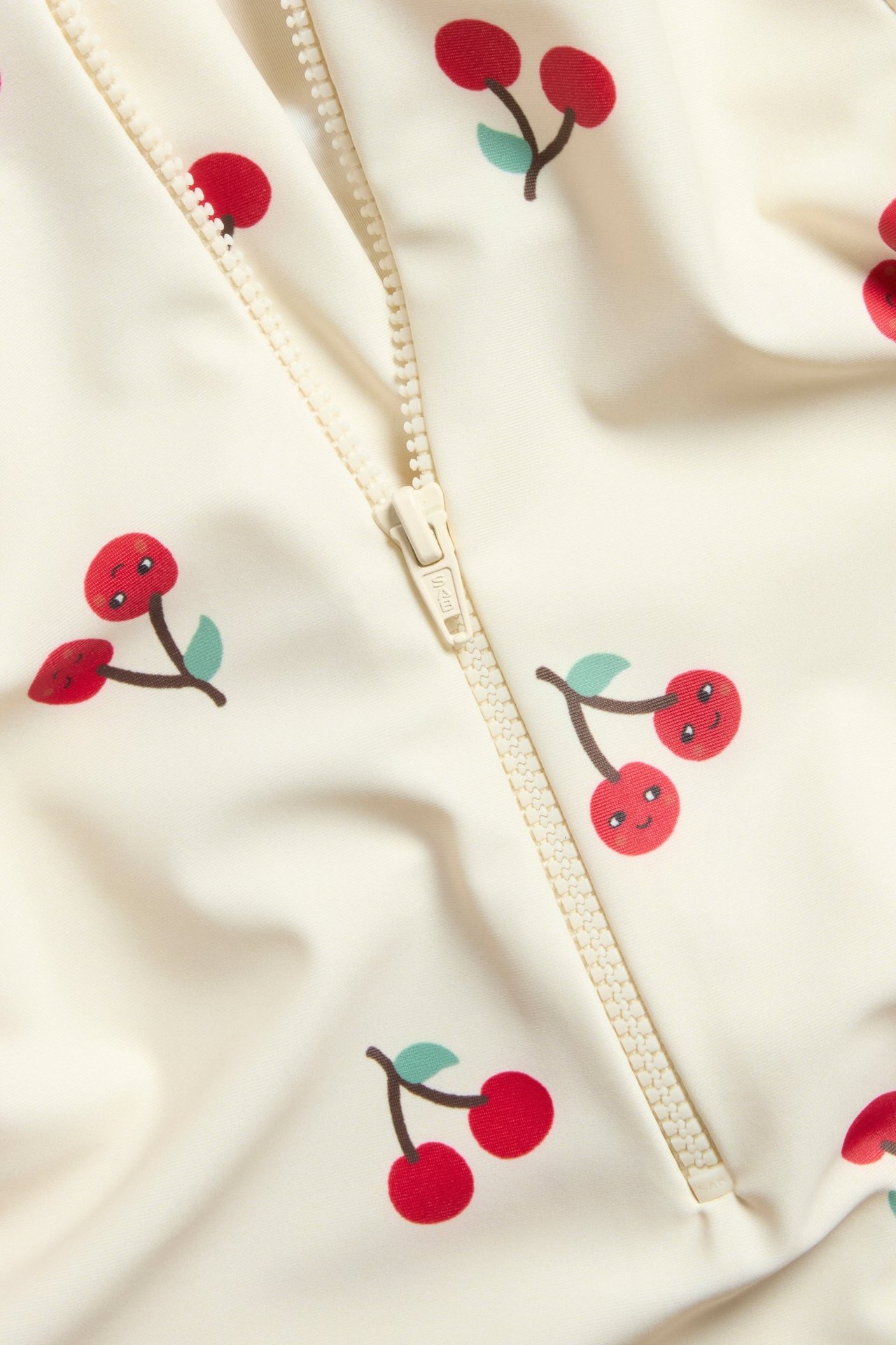 Närbild framifrån på en ljusbeige UV-dräkt med rött körsbärsmönster och dragkedja fram.
