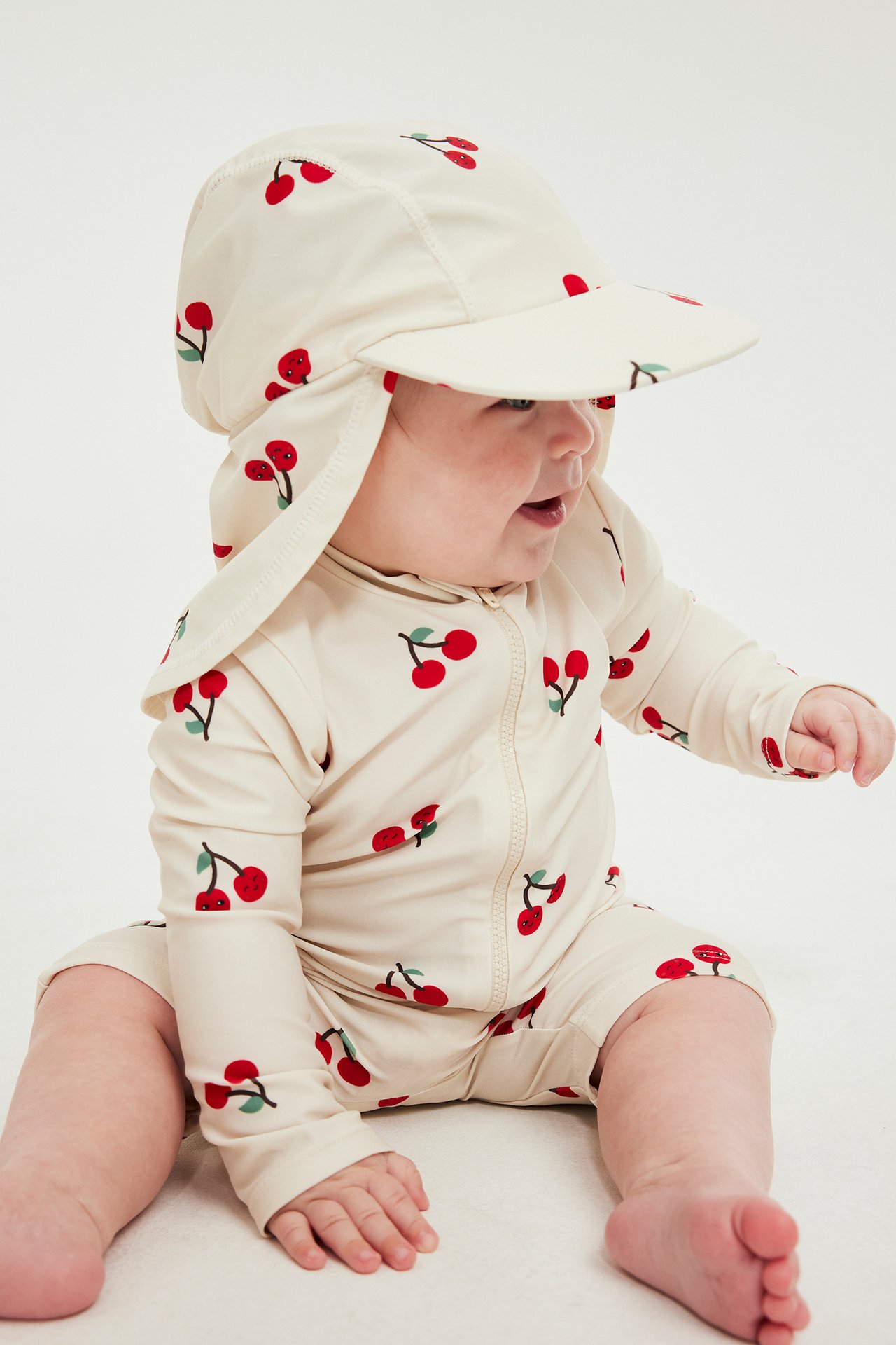 Sidebilde av en baby i lys beige UV-drakt med kirsebærtrykk og matchende UV-hatt.