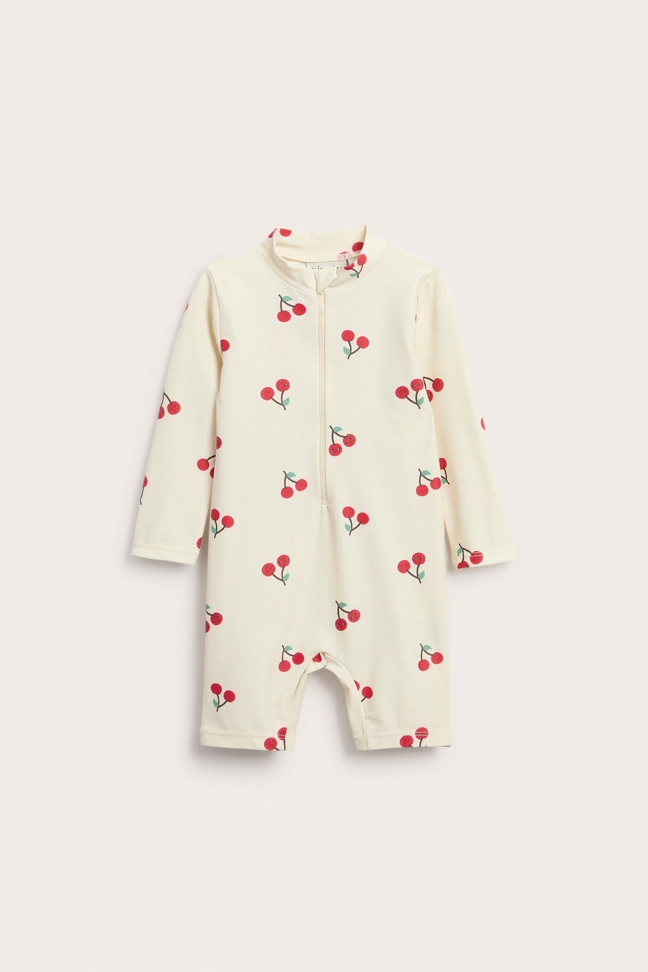 Frontvy av en baby UV-dräkt i ljusbeige med långa ärmar, dragkedja fram och rött körsbärstryck.