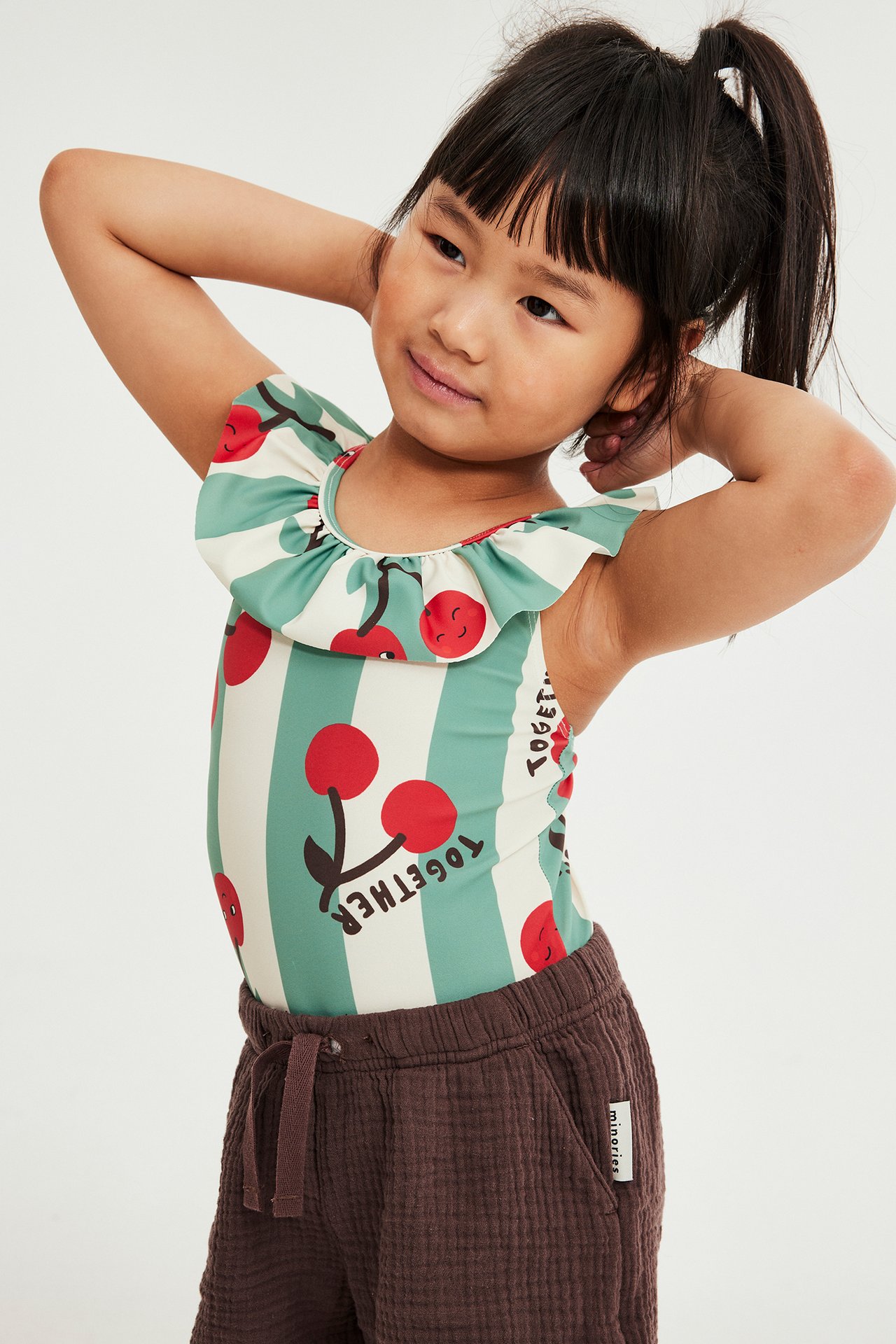 Barn, front-sida: beige/turkosrandig volangbaddräkt med körsbärsmönster, bärs med bruna shorts.