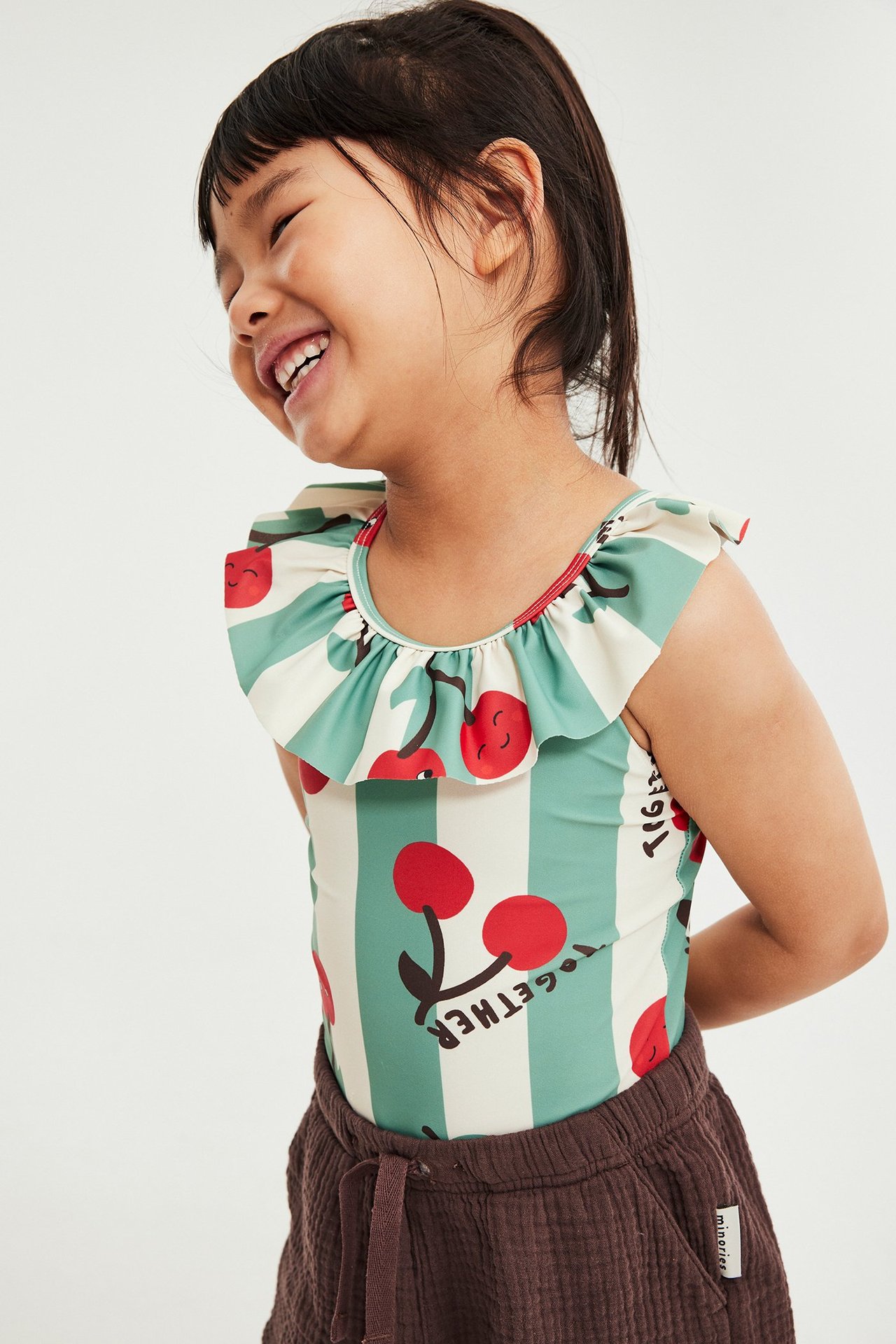 Sidovy av ett leende barn i randig ljusbeige och grön baddräkt med körsbärsmönster och volang, över mörkbruna shorts.