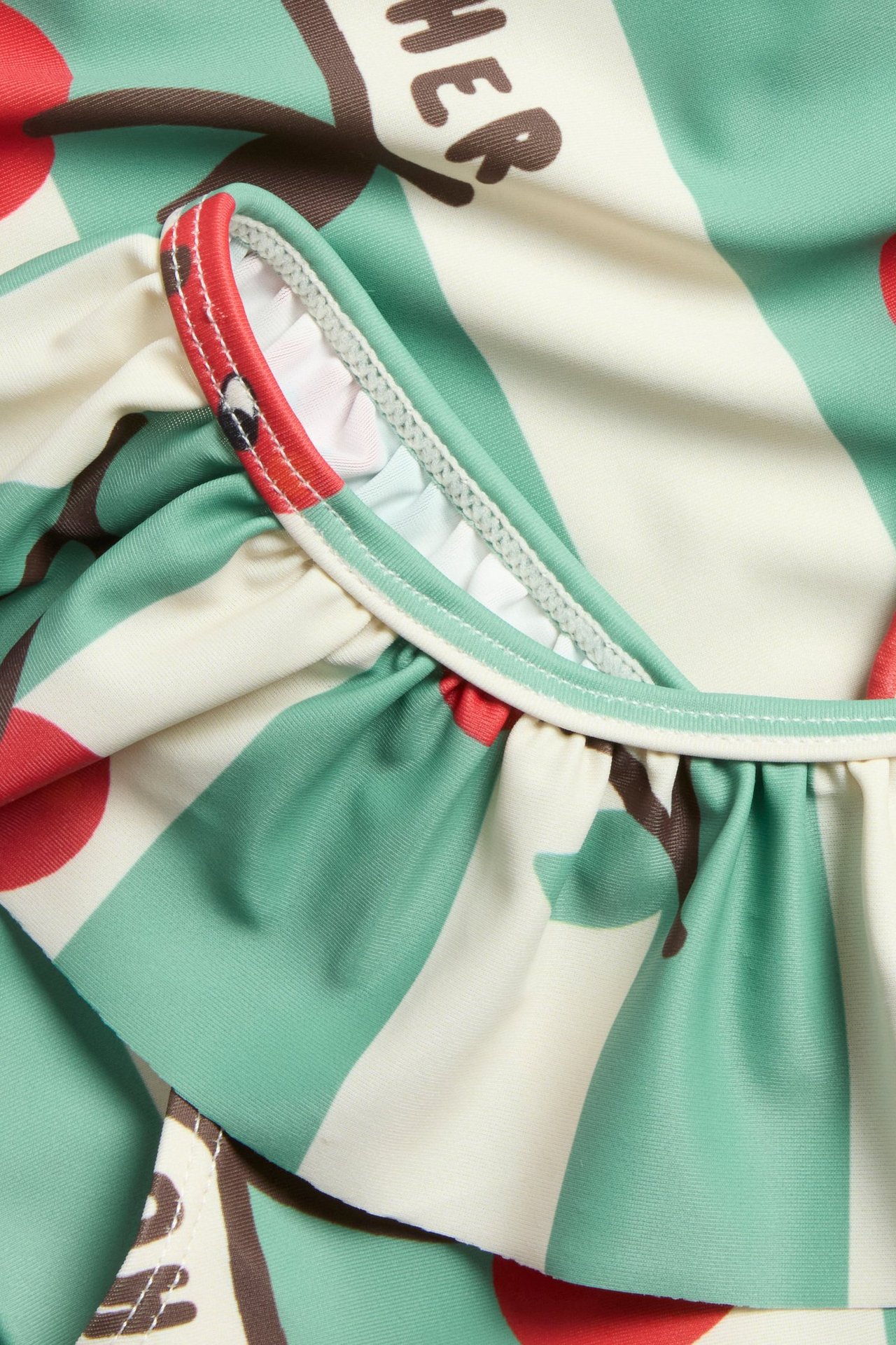 Närbild på ljus beige baddräkt med gröna ränder, rött körsbärsmönster och volangdetalj.