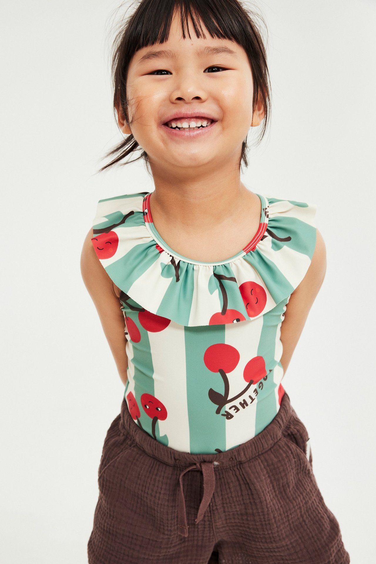 Przód, uśmiechnięte dziecko w jasnobeżowym kostiumie kąpielowym z falbanką i wzorem wiśni, na brązowych szortach.