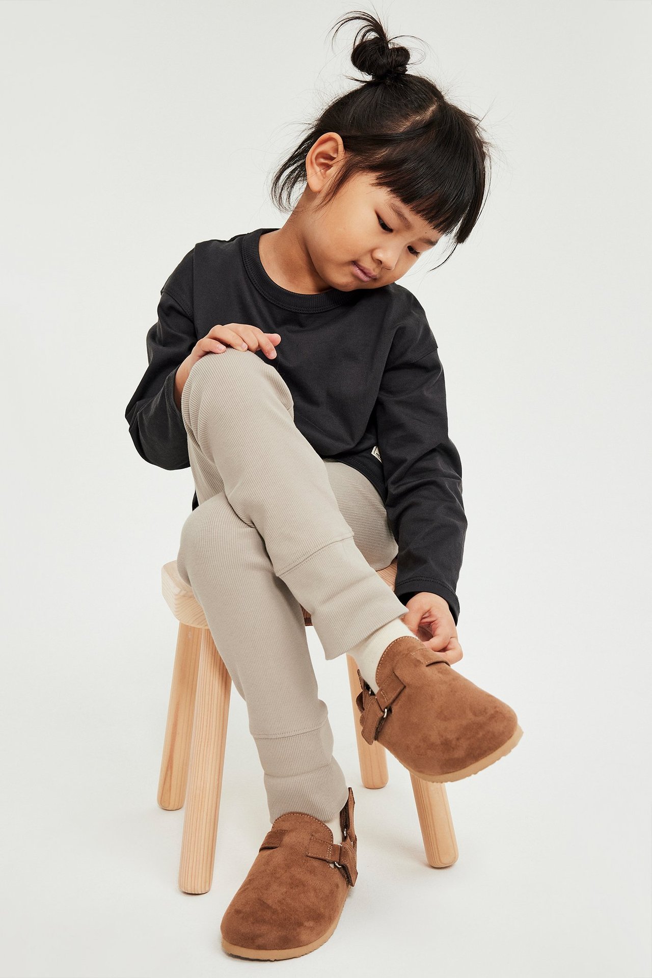 Dziecko w beżowych prążkowanych legginsach, czarnej koszulce z długim rękawem, białych skarpetkach i brązowych butach.