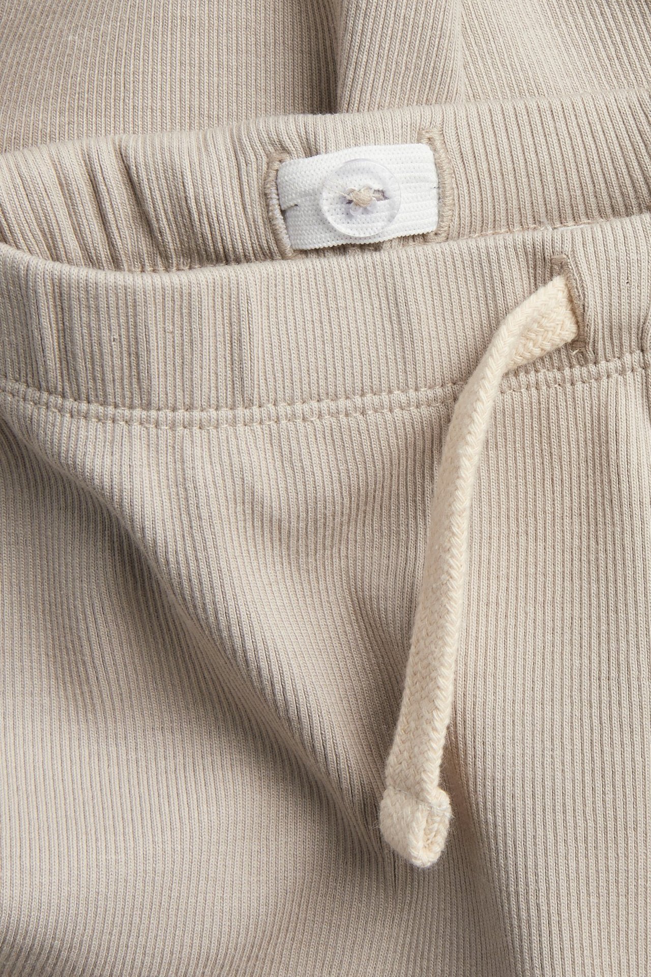 Zbliżenie beżowych prążkowanych legginsów w pasie z regulowaną gumką na guziki i ozdobnym sznurkiem.