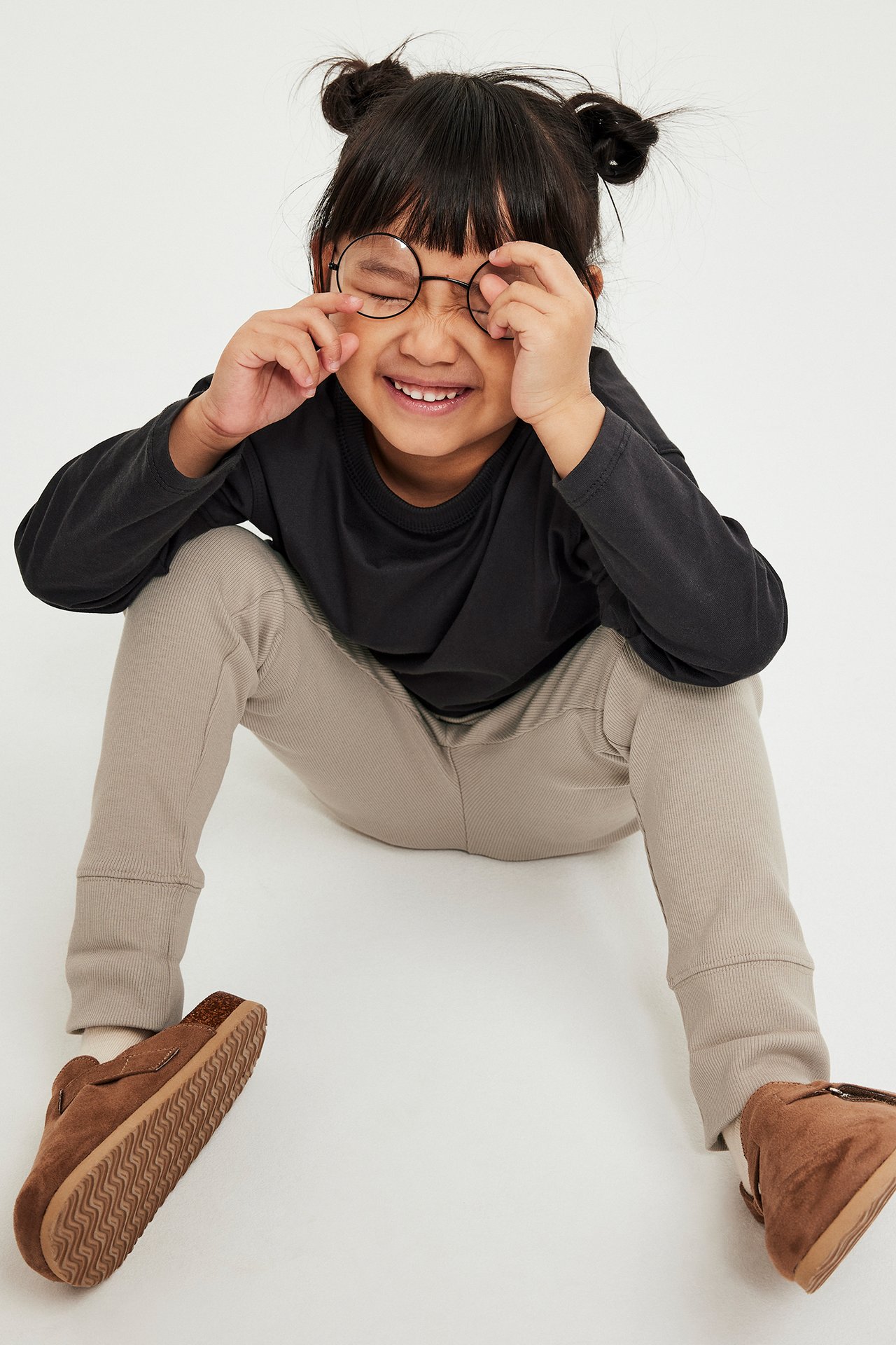 Widok z przodu: Dziecko w beżowych prążkowanych legginsach z wywijanymi mankietami, czarnej bluzce i brązowych kapciach.