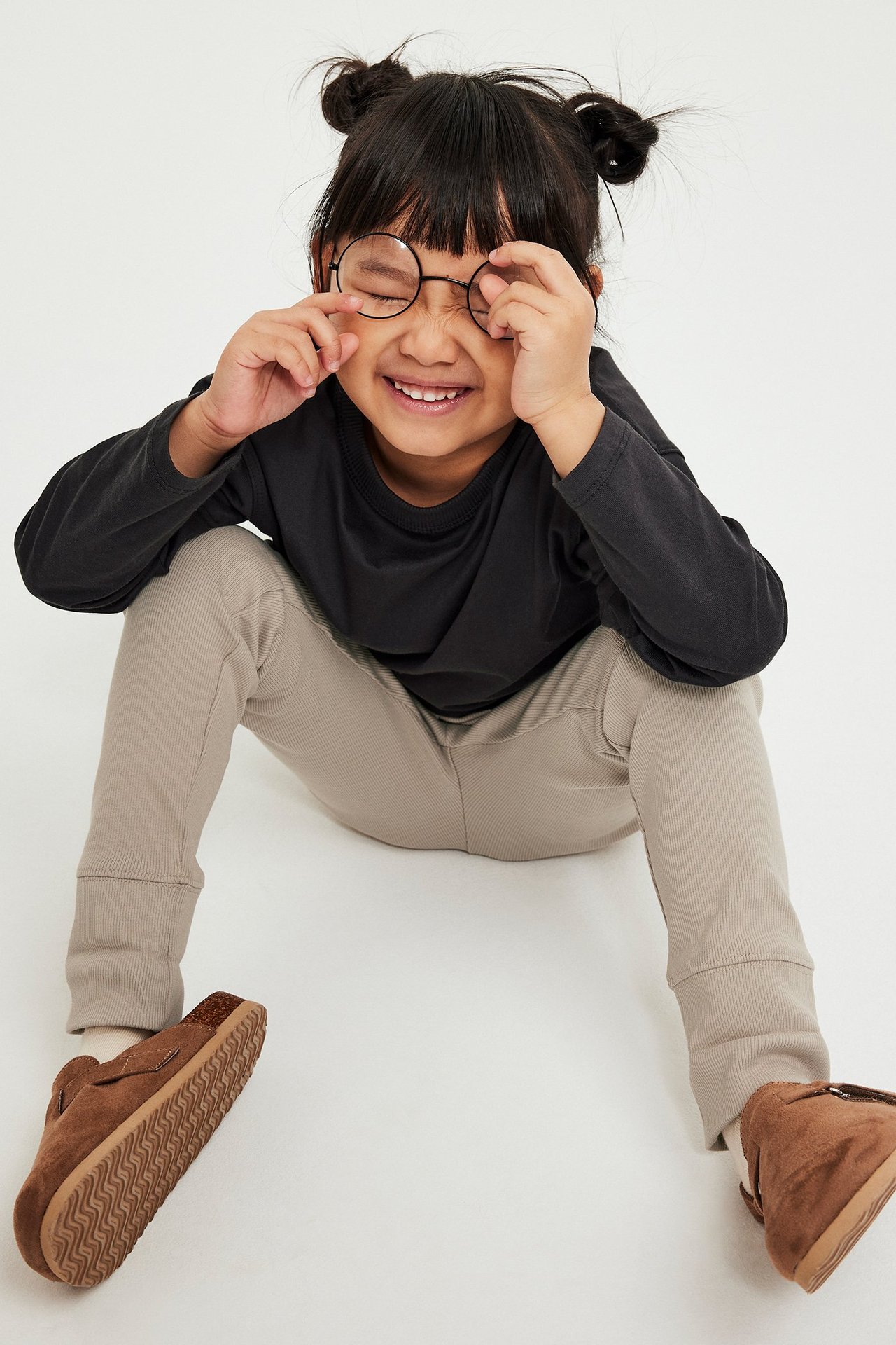 Widok z przodu: Dziecko w beżowych prążkowanych legginsach z wywijanymi mankietami, czarnej bluzce i brązowych kapciach.