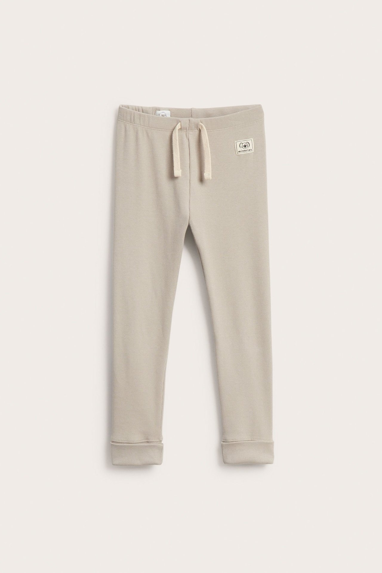 Widok z przodu beżowych prążkowanych legginsów dziecięcych z ozdobnym białym sznurkiem i wywijanymi mankietami.