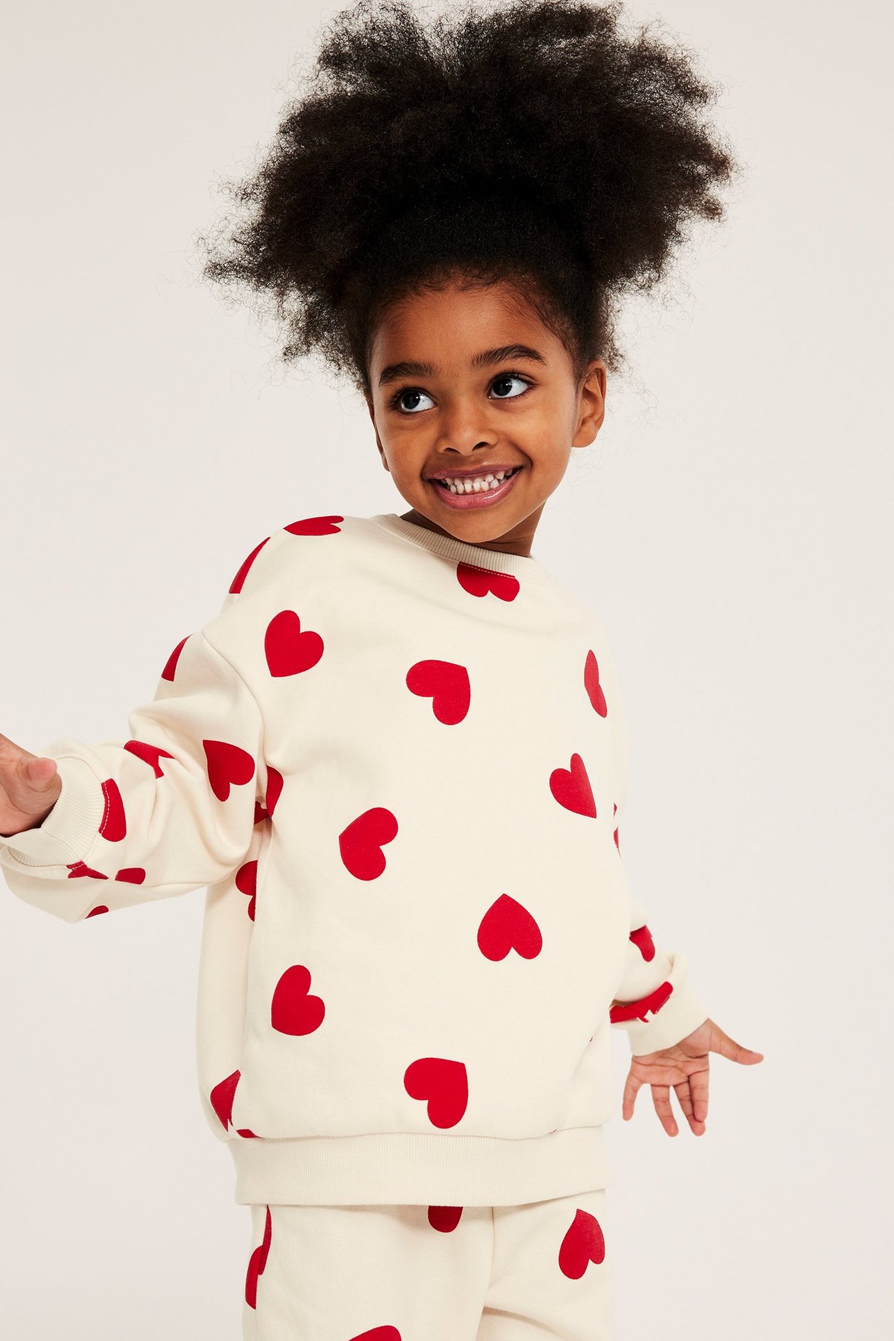Widok z przodu dziecka w off-white, szczotkowanej bluzie z czerwonymi sercami i pasującymi spodniami.