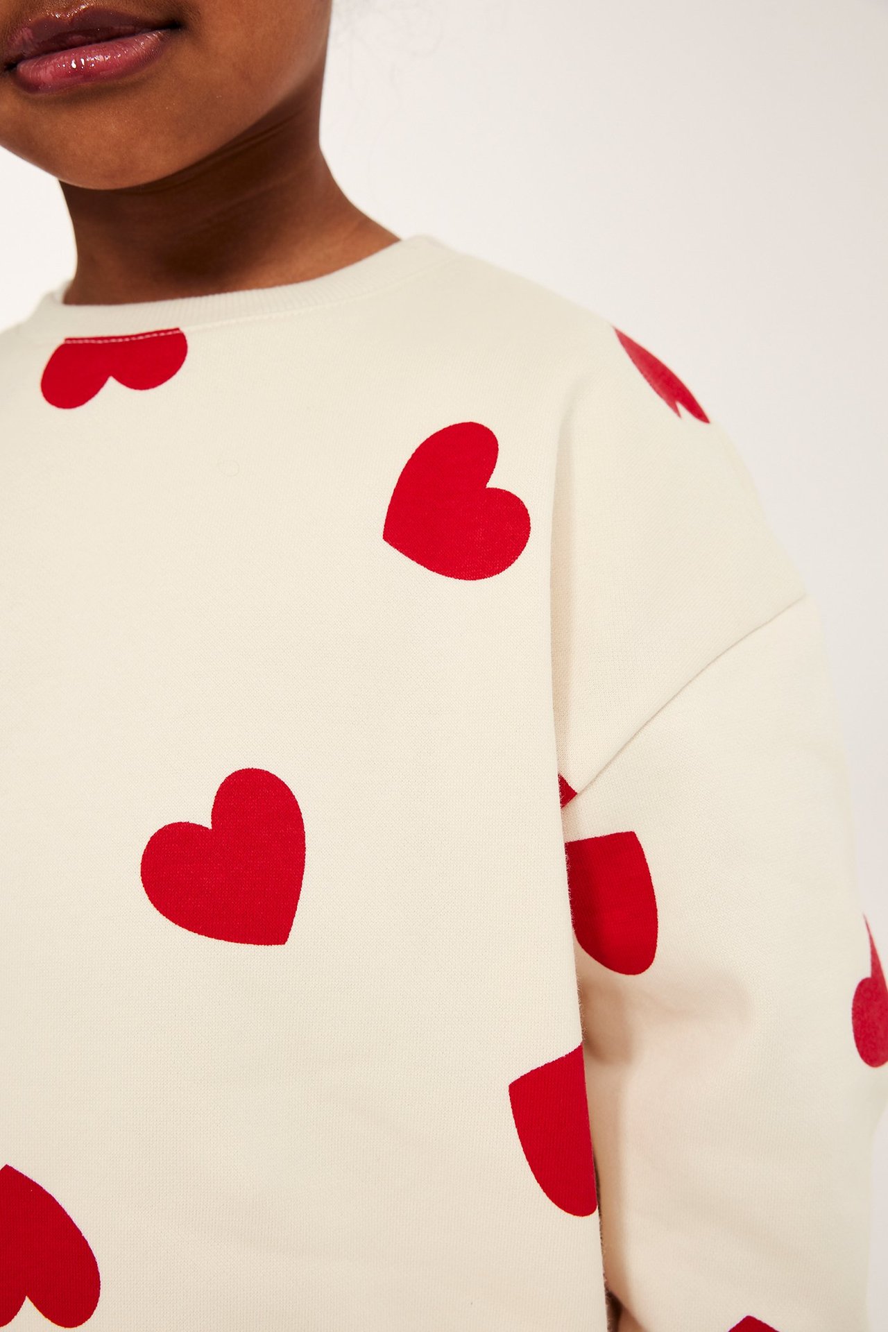 Zbliżenie dziecka w ecru szczotkowanej bluzie z nadrukiem czerwonych serc na całej powierzchni.