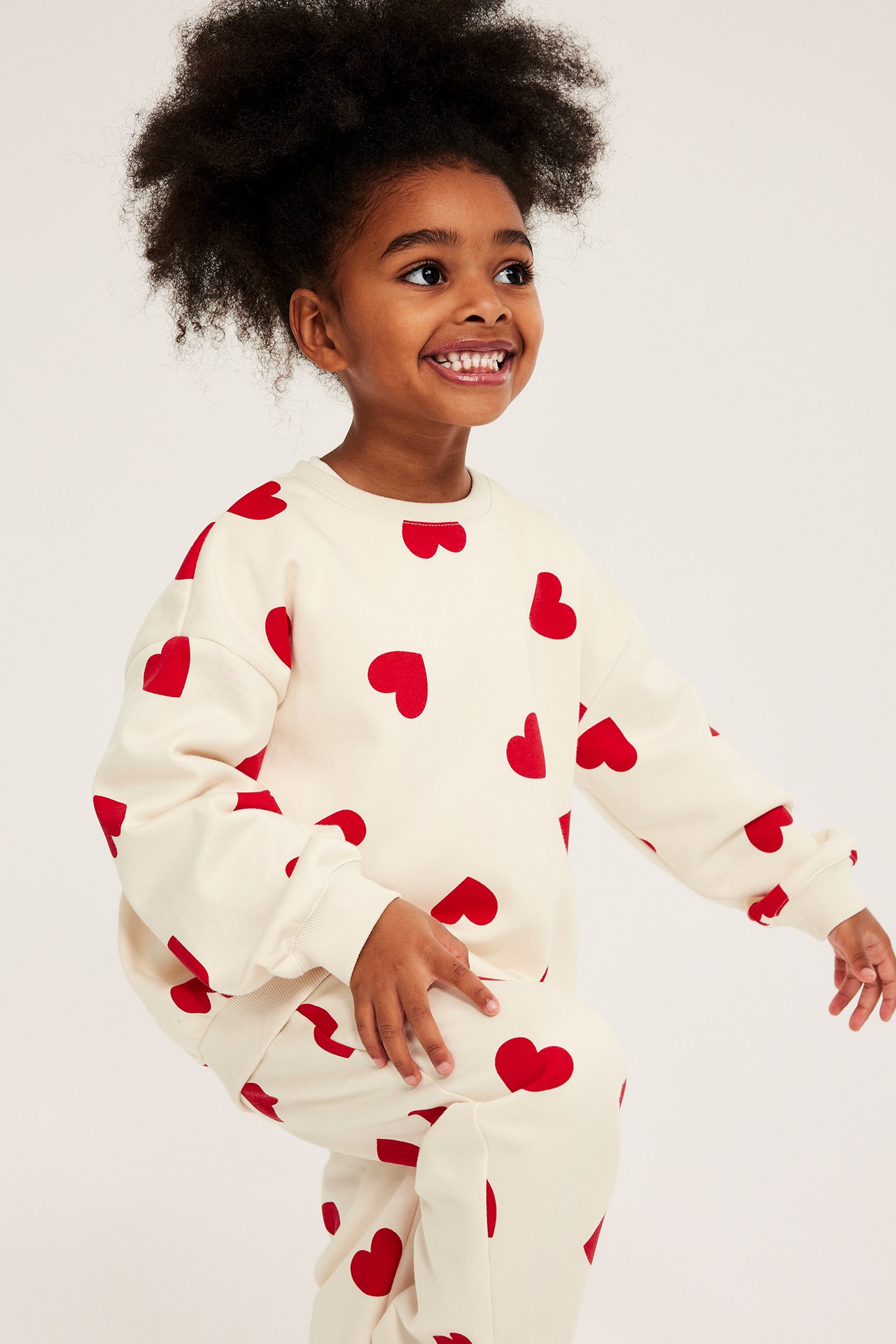 Uśmiechnięte dziecko w ecru bluzie i pasujących spodniach dresowych z czerwonymi sercami.