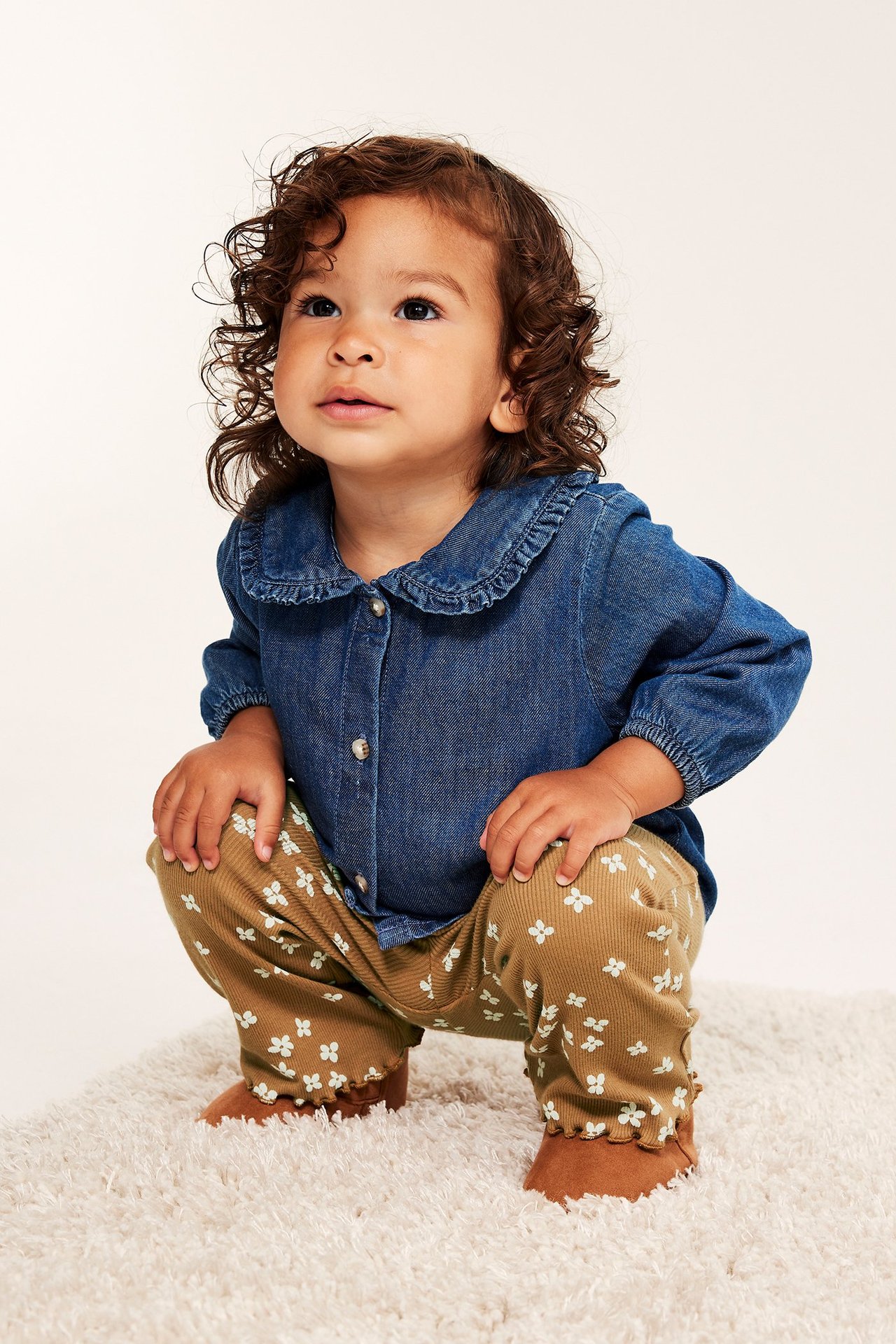 Forfra: baby i brune, blomstrete, ribbede flared-leggings og en blå dongeriskjorte.