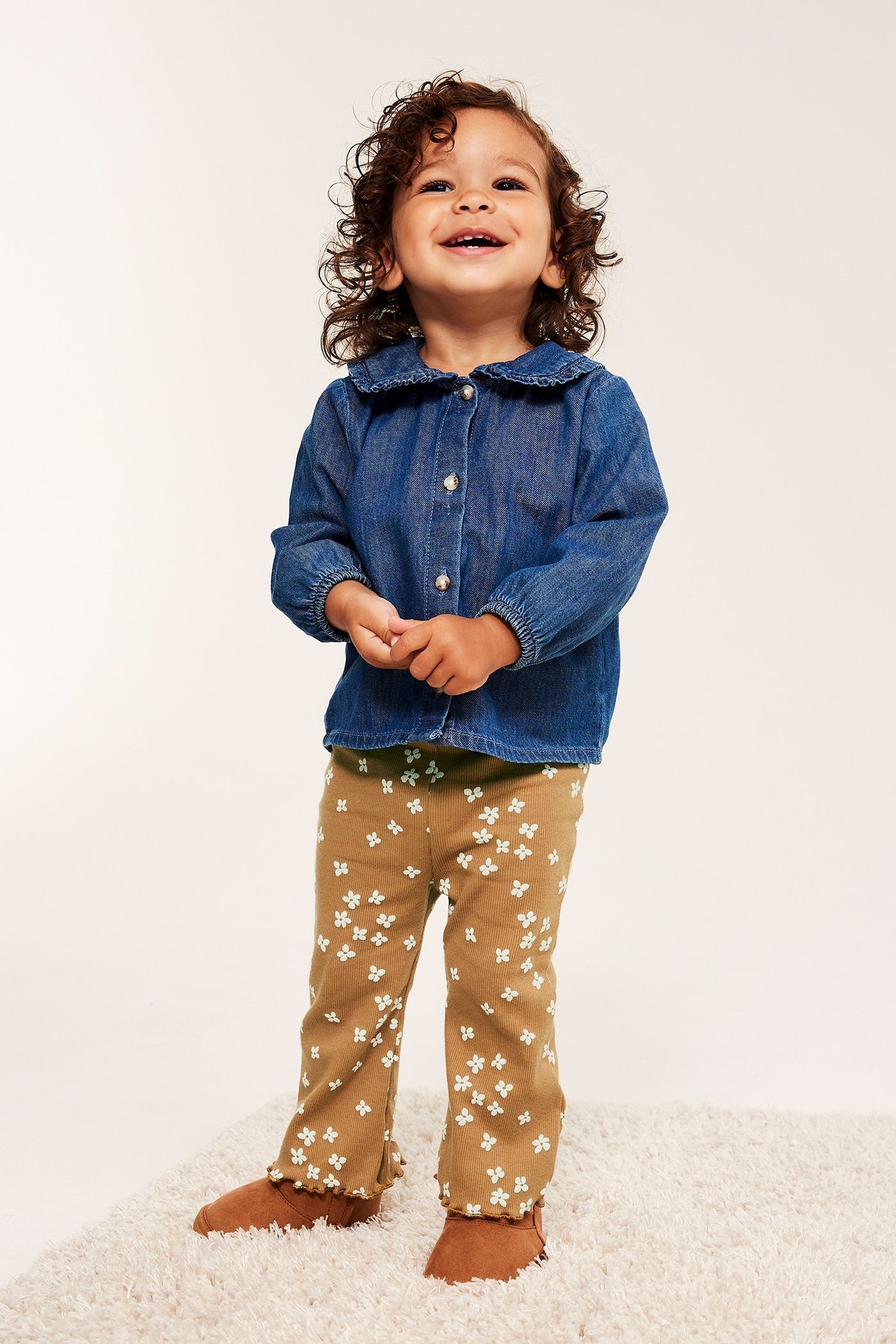 Forfra, en baby har på seg brune blomstrete flare-leggings, en blå dongeriskjorte og brune sko.
