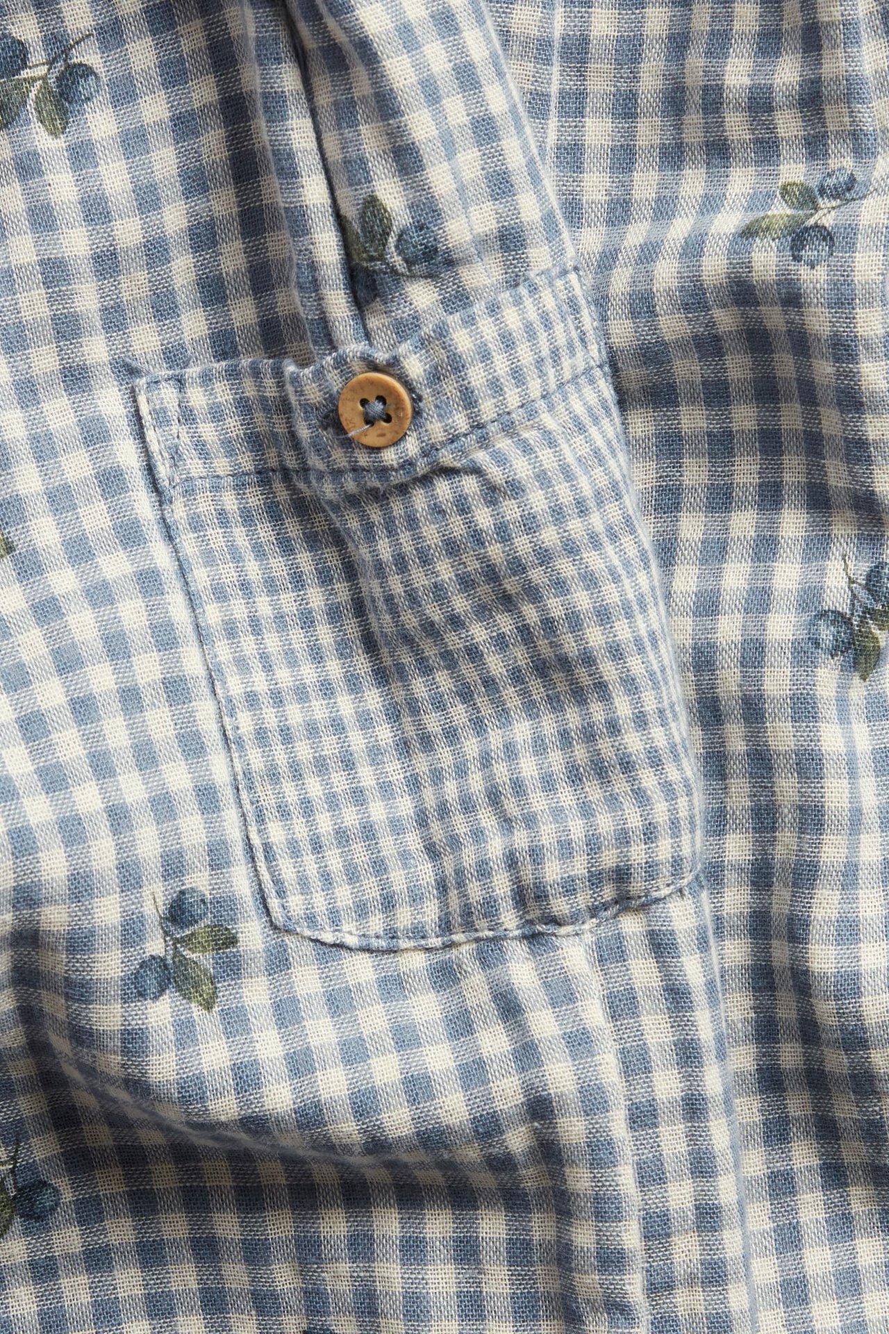 Nærbilde: Blå- og hvitrutet bomullsstoff med blåbærmønster, en påsydd lomme og treknapp.