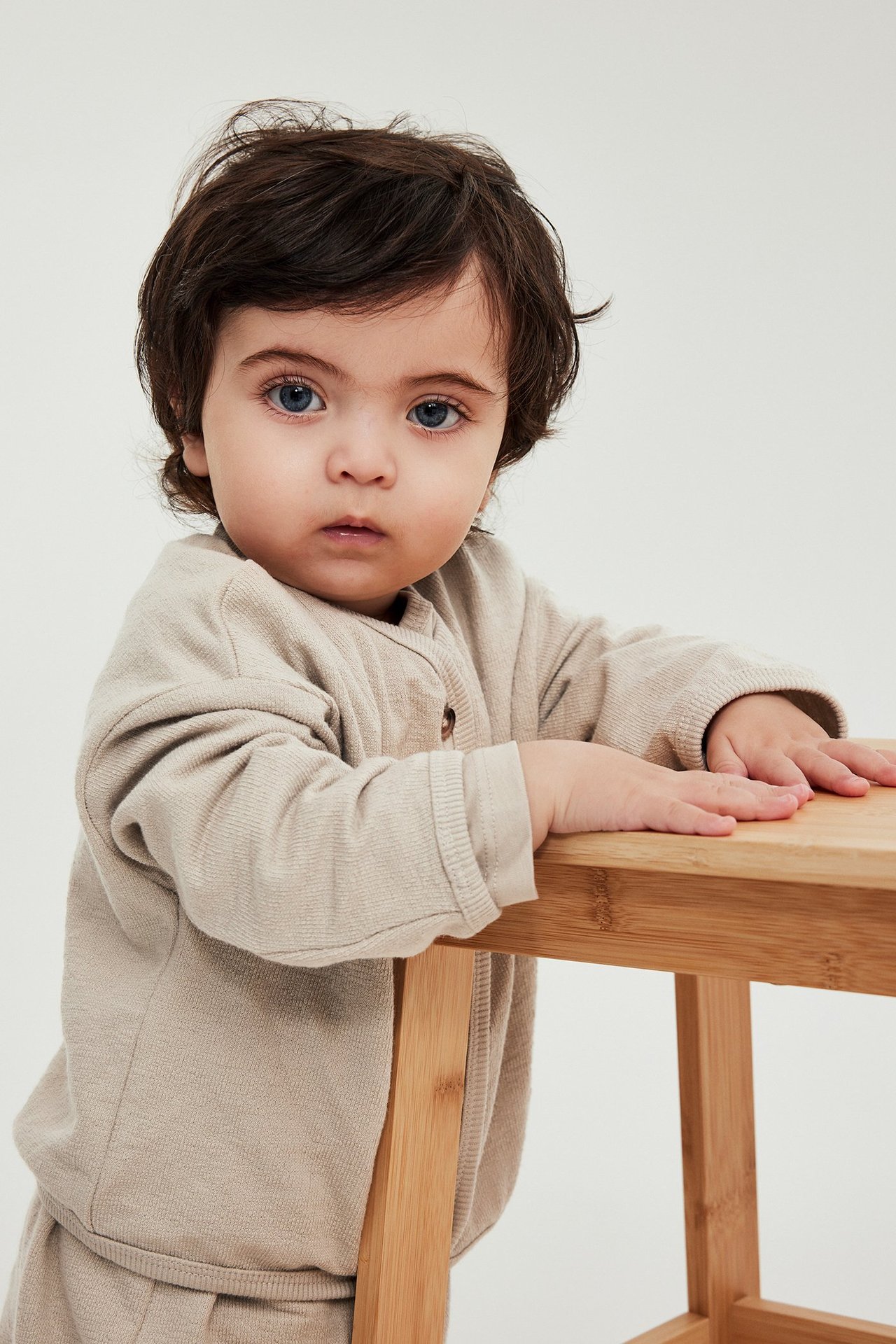 Edestä: vauva pukeutunut beigeen napilliseen puuvillaneuletakkiin, pitää kiinni puisesta jakkarasta.