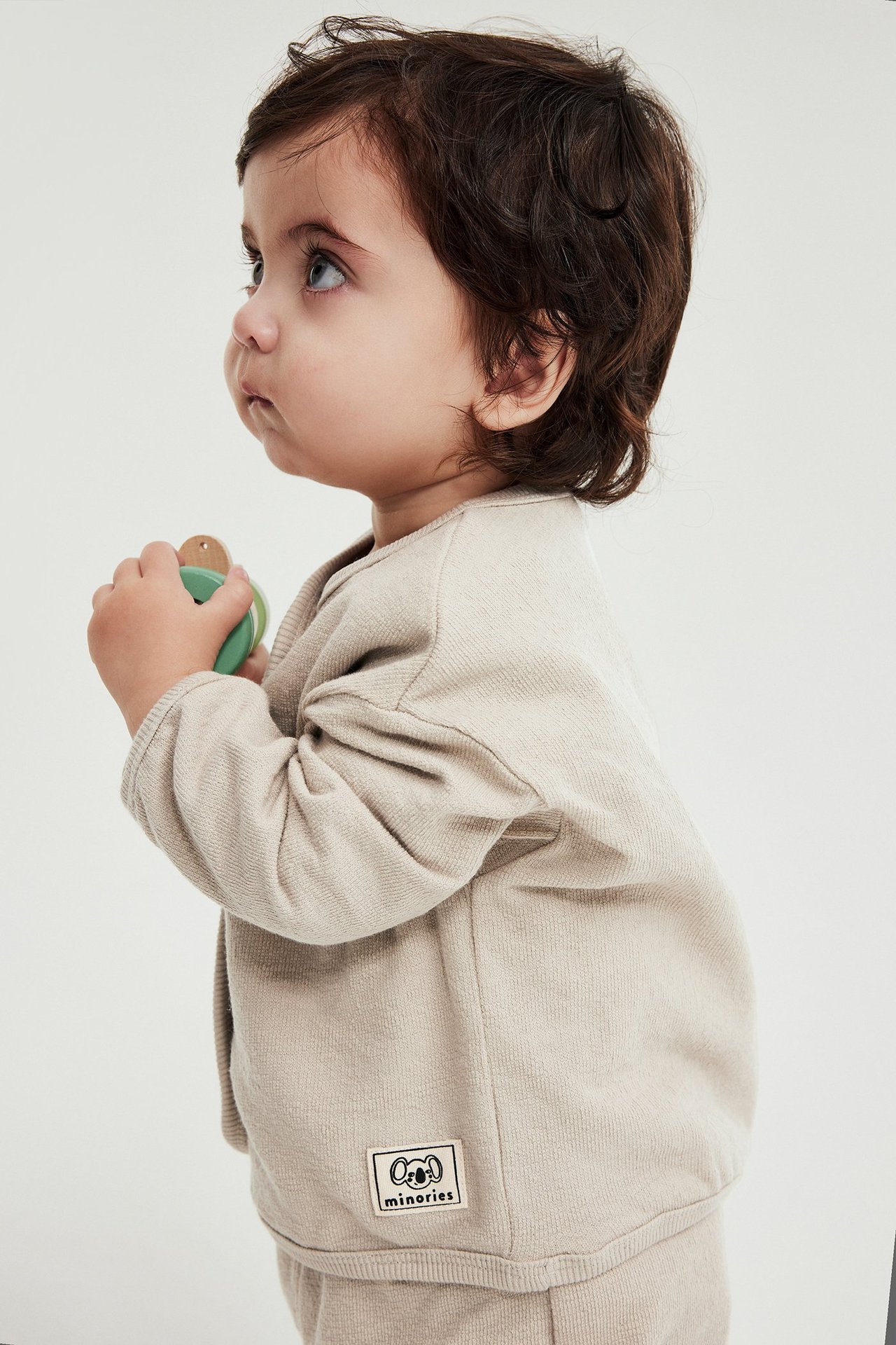 Sivukuva vauvasta beigessä puuvillaneuletakissa ja yhteensopivissa housuissa, pitelee vihreää lelua.